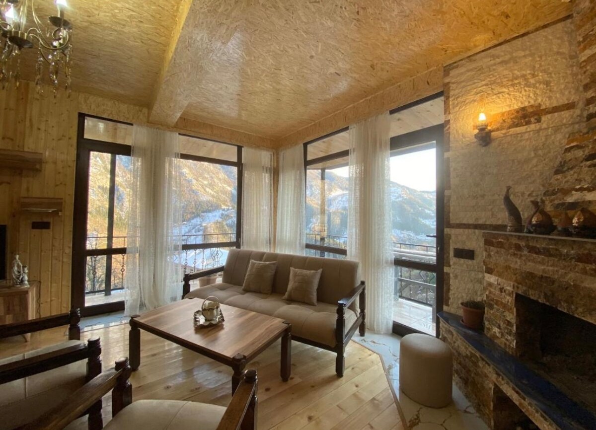A cozy living area features large windows framing views of the surrounding mountains. Comfortable seating arrangements include two armchairs and a sofa. Natural materials like wood and stone are used throughout, while soft drapes partially filter the light, creating a warm and inviting space.