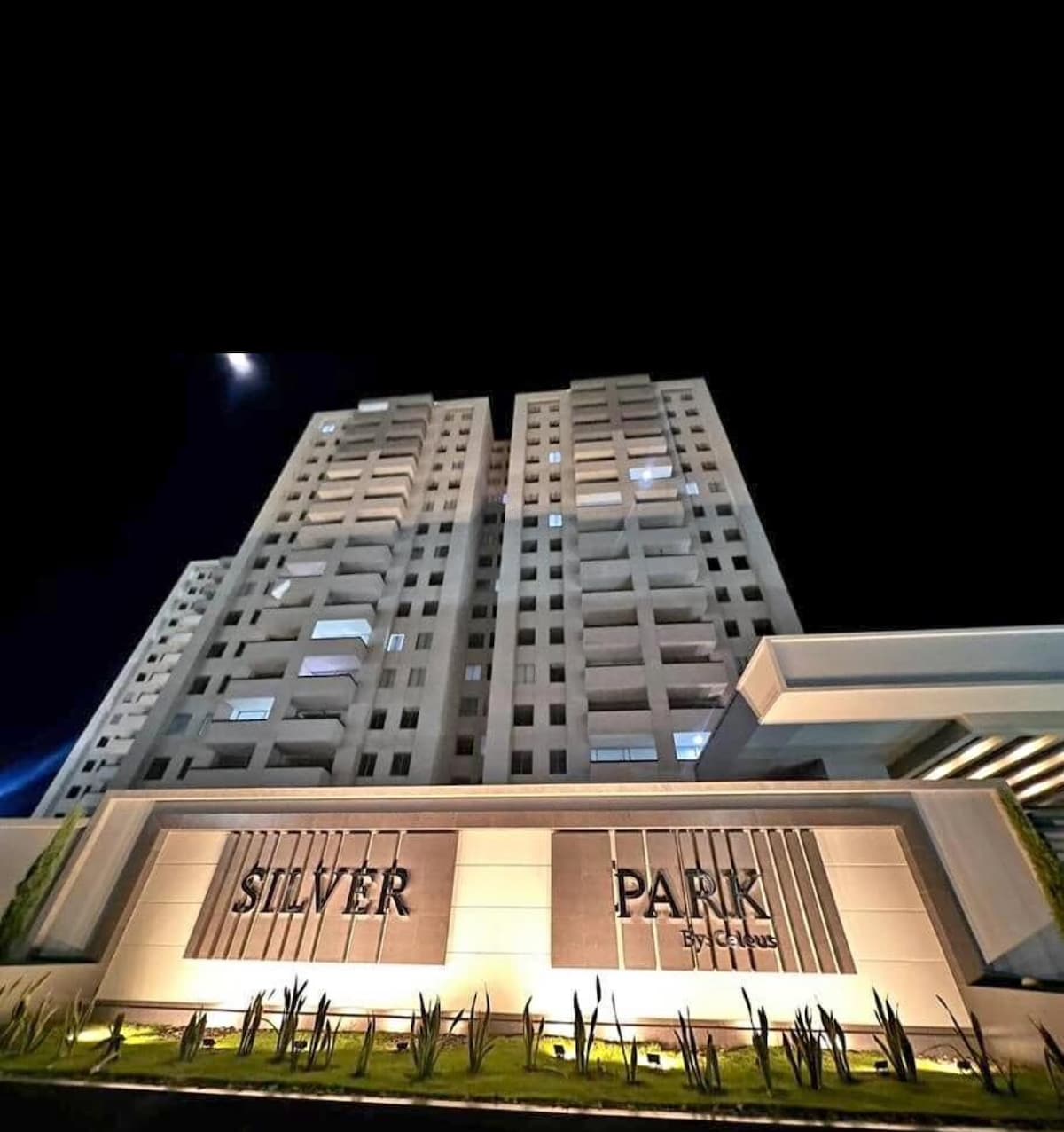 The exterior of the Silver Park condominium is displayed, featuring a modern design with a well-lit entrance. Elevated floors are visible, accompanied by a landscaped area with greenery. The building’s name is prominently featured on a sleek sign, illuminated against the night sky.
