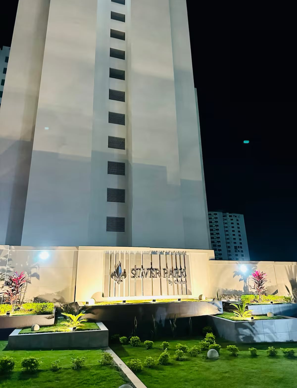 The image captures the exterior of the Silver Park condominium building at night. Soft lighting illuminates the entrance, enhancing the lush greenery surrounding the well-maintained landscape. The building's modern architecture and prominent signage are clearly visible against the night sky.