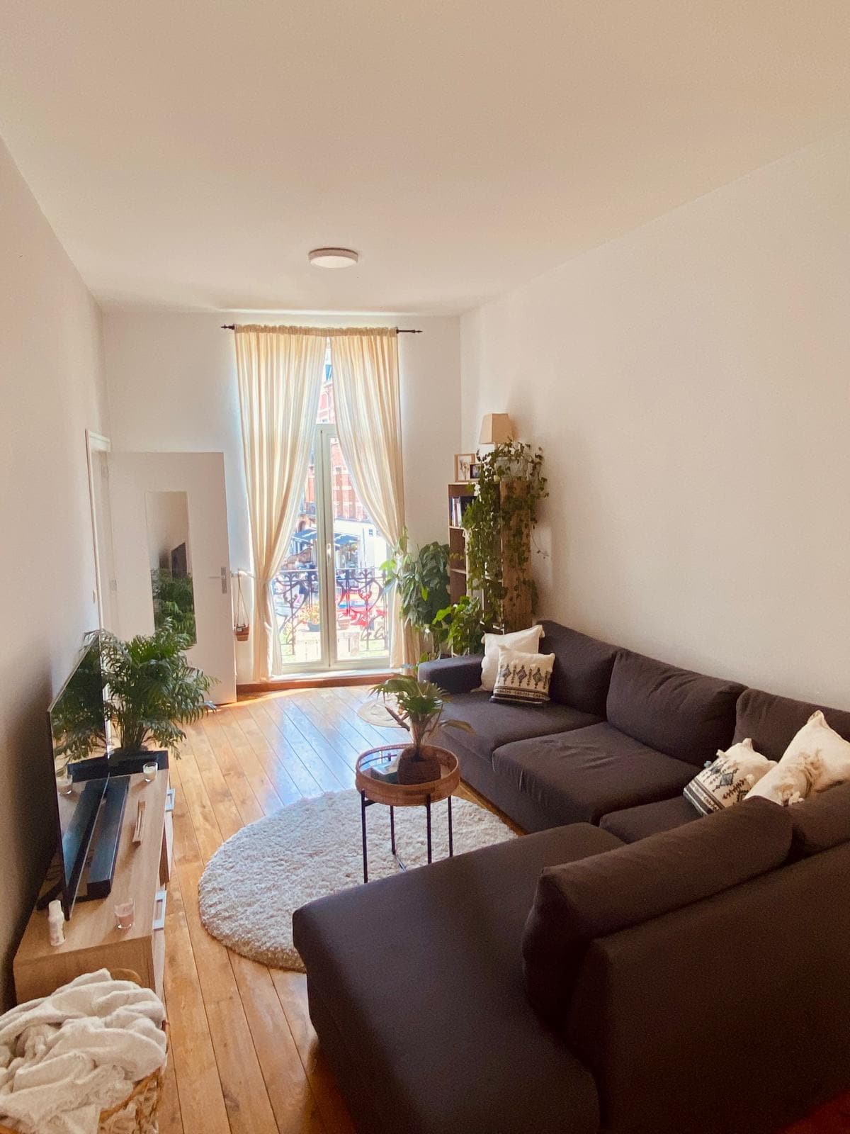 A bright living area features a spacious dark sofa inviting relaxation. A round coffee table is positioned in front of the sofa, while greenery adds a touch of nature. Natural light enters through large glass doors leading to a balcony, enhancing the open feel of the space.