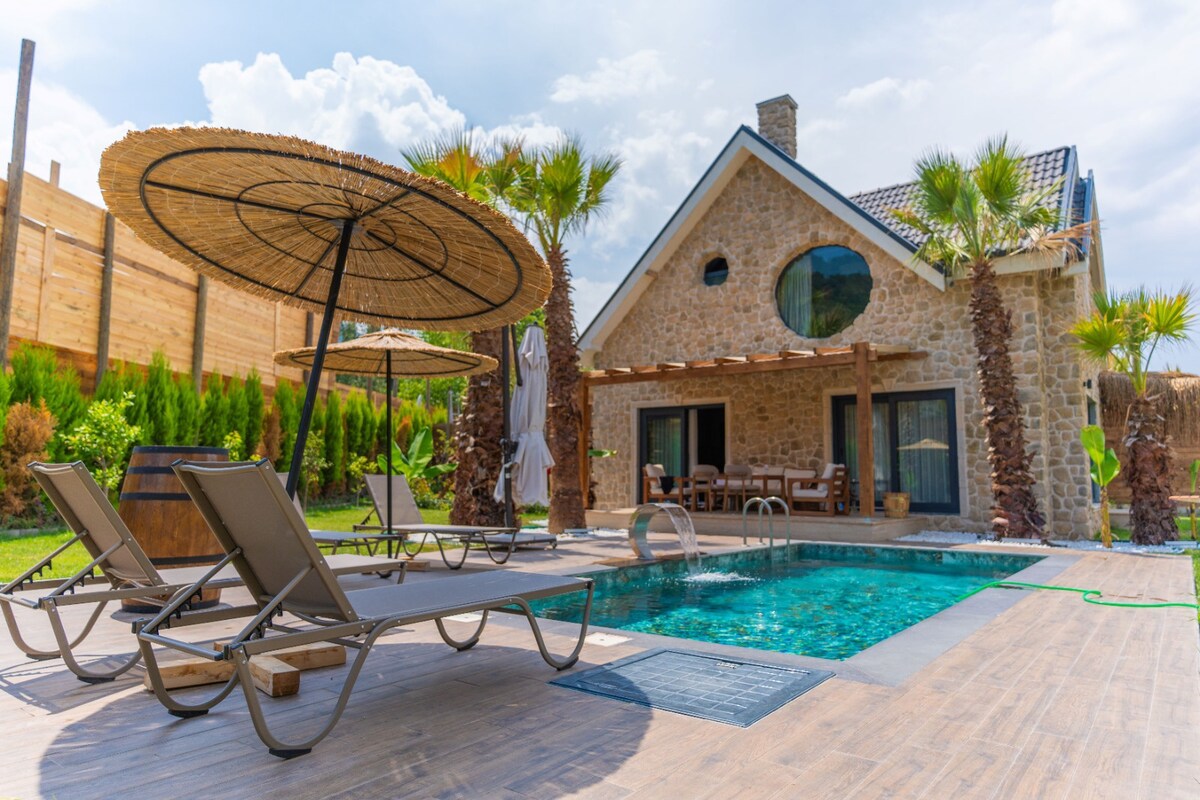 A stone house with a round window is framed by lush palm trees and features a private pool. Poolside, two loungers are sheltered under large umbrellas. A wooden dining table and chairs are visible in the outdoor area, surrounded by greenery.