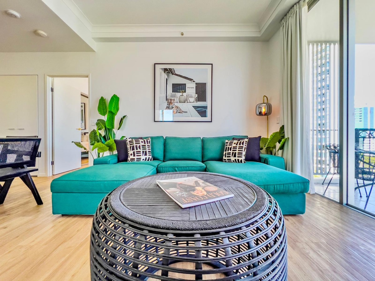 A modern living area features a spacious turquoise sofa adorned with patterned cushions. A circular woven coffee table displays a magazine. Lush green plants are positioned nearby, and large glass doors provide access to a balcony filled with natural light.