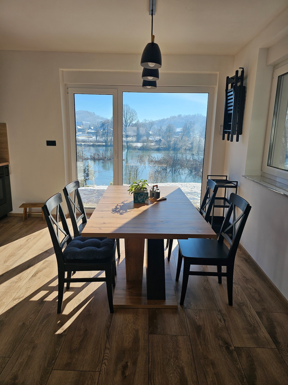 A dining area features a wooden table surrounded by six black chairs, with a small potted plant at the center. Large sliding glass doors offer a view of the river and landscape beyond, creating a bright and inviting space.