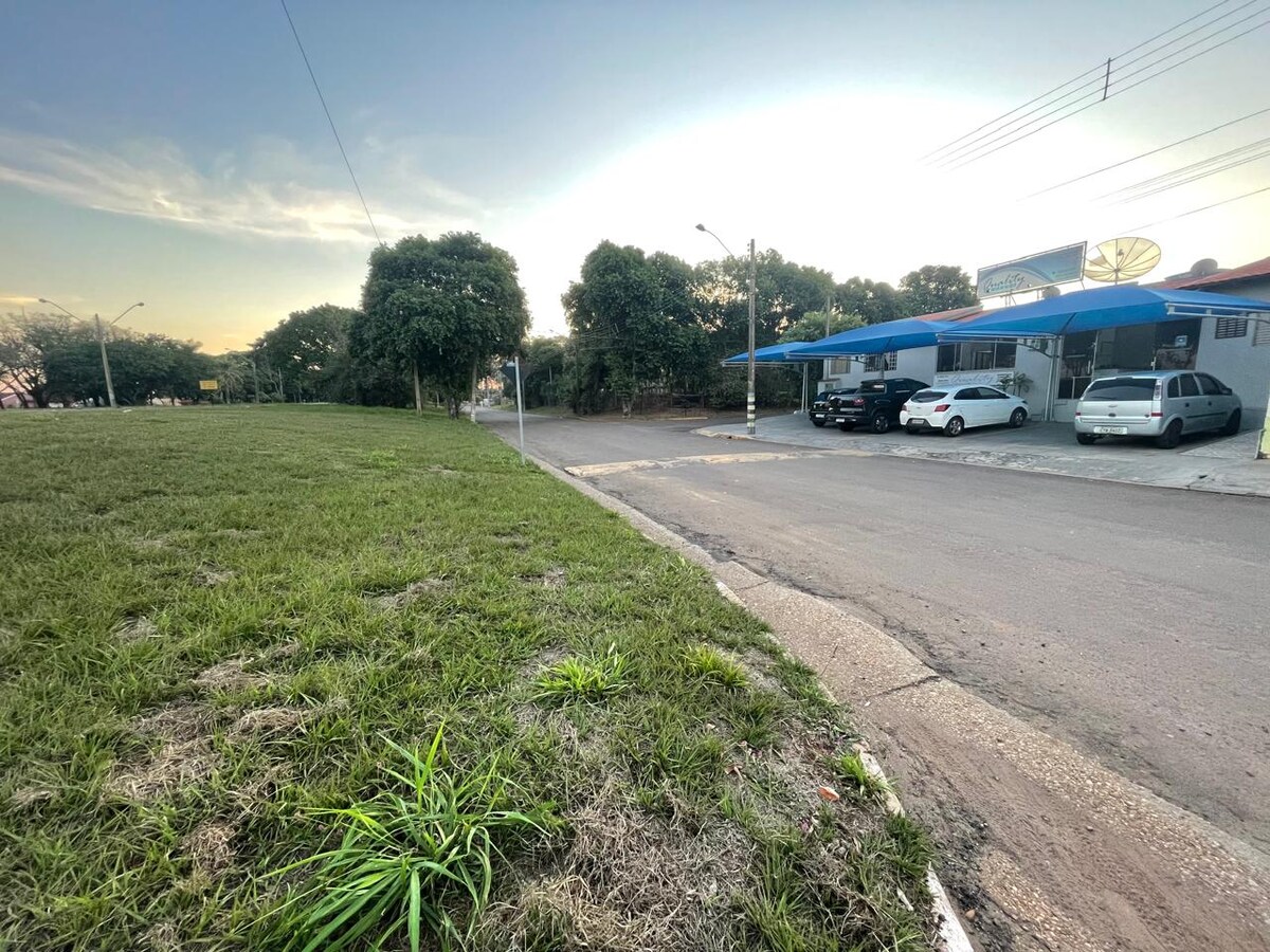 A view of the neighborhood shows a grassy area leading to a paved street. Vehicles are parked along the curb near a building with a blue awning. Trees line the street, contributing to a spacious and open environment.