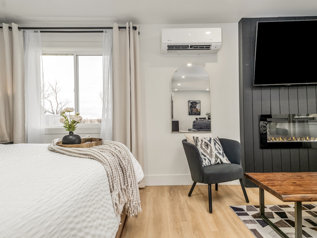 A cozy bedroom setting is presented with a spacious bed dressed in a textured blanket. A large mirror reflects light from the window, while a small table holds a decorative tray and a potted plant. A wall-mounted television and electric fireplace add modern touches to the space.