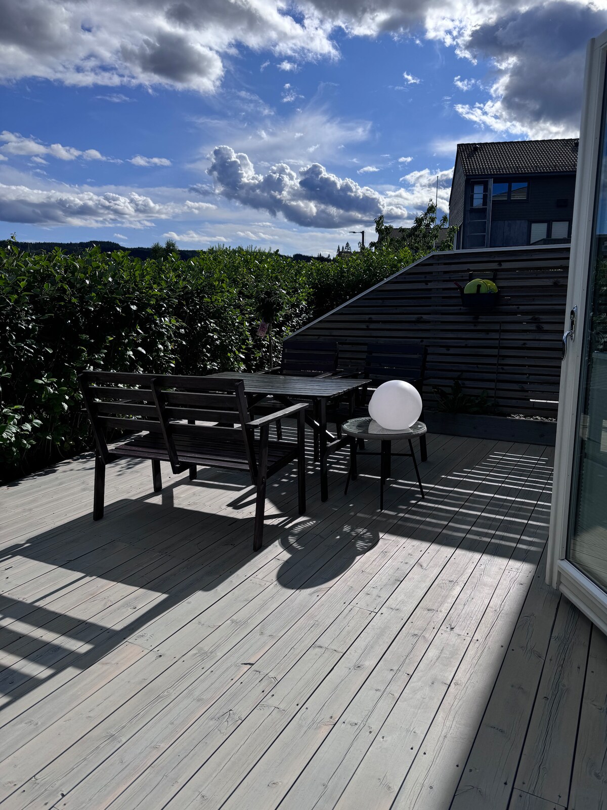 An outdoor terrace features a dining table with four chairs and a small side table. Soft shadows are cast on the wooden deck by the furniture. Lush greenery surrounds the space, and a clear sky with fluffy clouds is visible above.