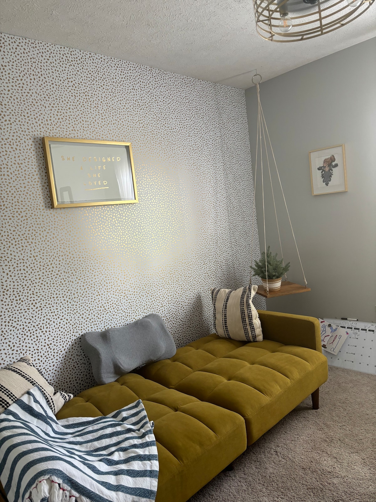 A cozy seating area features a mustard-colored tufted couch against a dotted wallpaper. Two decorative pillows add texture, while a small potted plant sits on a nearby wooden table. Light is gently filtered through a large window, illuminating the space.
