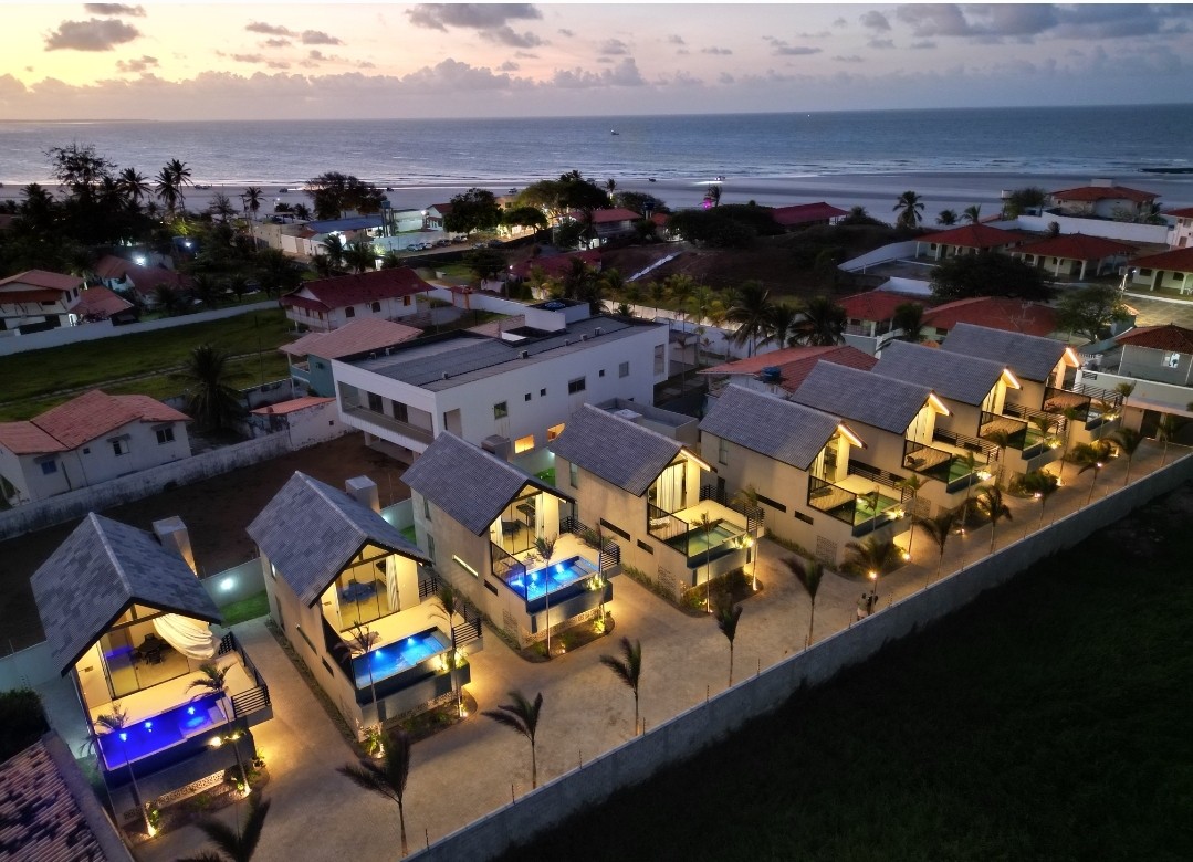 An aerial view displays a series of two-story cabins illuminated by soft lighting, arranged in a line. Each cabin features a small private pool on the balcony, with a backdrop of the ocean and nearby residential areas under a twilight sky.