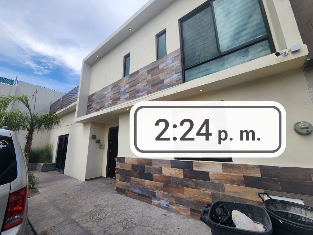 The exterior of a modern building features a mix of textured stone and smooth surface finishes. Large windows allow for natural light, while palm trees frame the entrance. A vehicle is parked in front, and a decorative trash bin is positioned to the side.