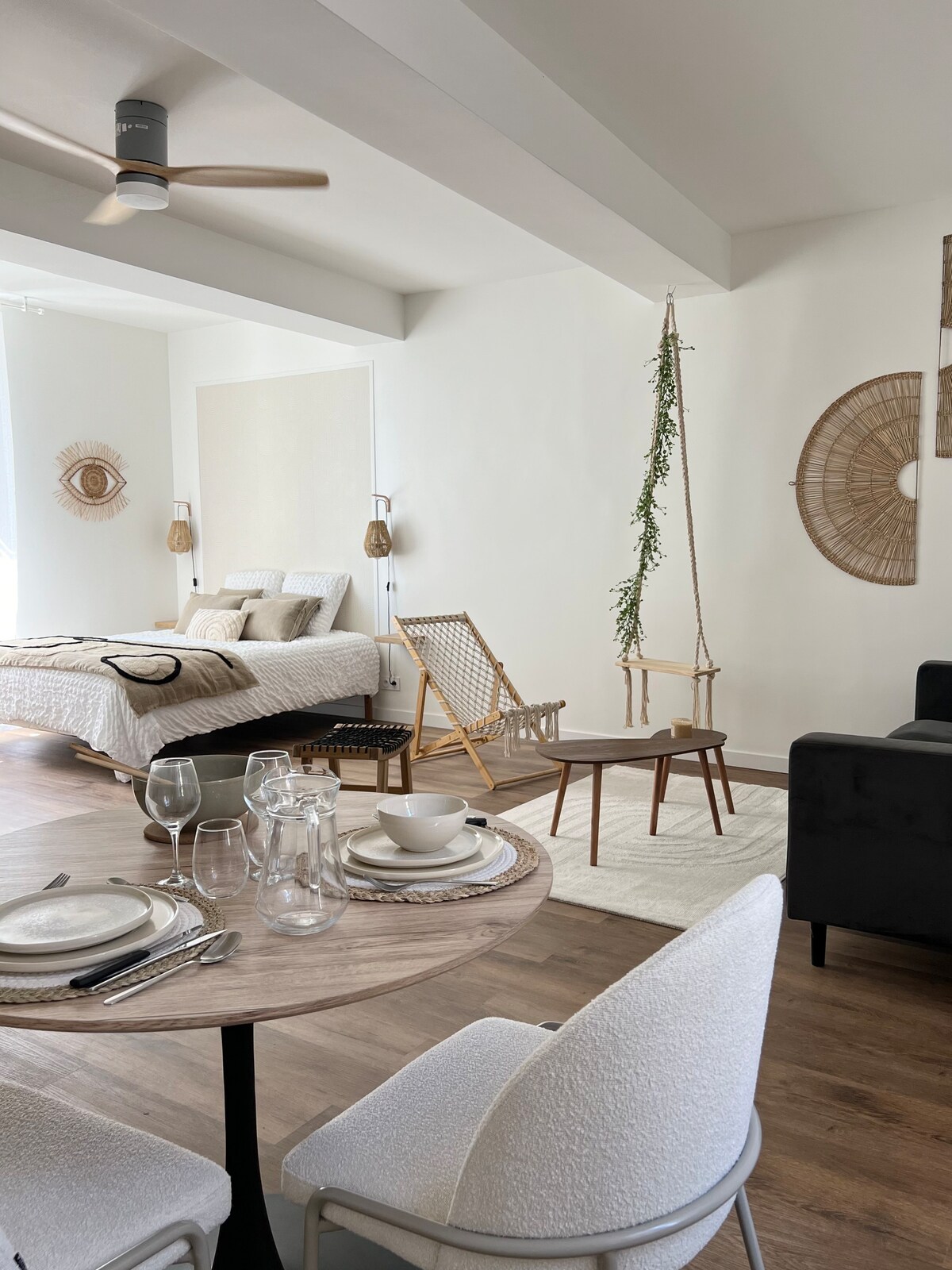A well-organized living space features a dining area with a round table and stylish chairs. Nearby, a cozy bedroom area includes a spacious bed, complemented by decorative elements such as a hanging chair and wall art. Natural light enhances the inviting atmosphere.