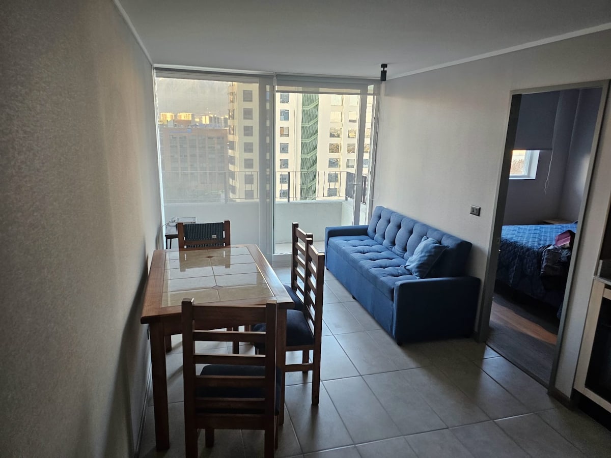 The living and dining area is furnished with a wooden table and four chairs, complemented by a blue futon. Large windows allow natural light to fill the space, providing views of the city skyline.