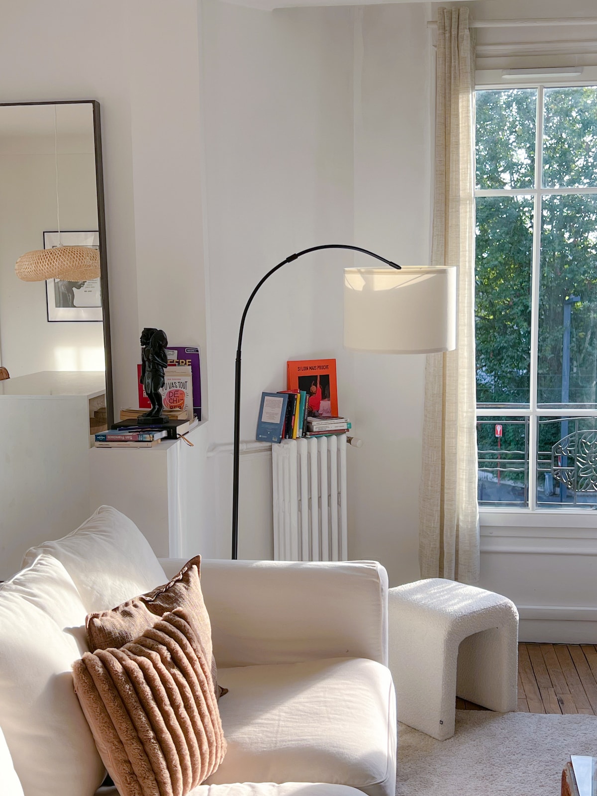 A well-lit living area is presented with a white sofa adorned with plush pillows. A minimalist floor lamp stands next to the sofa, and a decorative table is positioned nearby. A large window allows natural light to enter, framing the view of greenery outside.