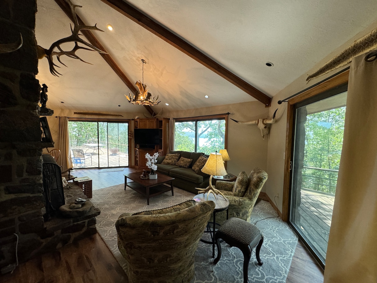 A spacious living area features large glass doors offering views of surrounding trees and outdoor space. Comfortable seating is arranged around a coffee table, with a stone fireplace adding a rustic touch. Decorative antlers and a chandelier enhance the cozy ambiance.