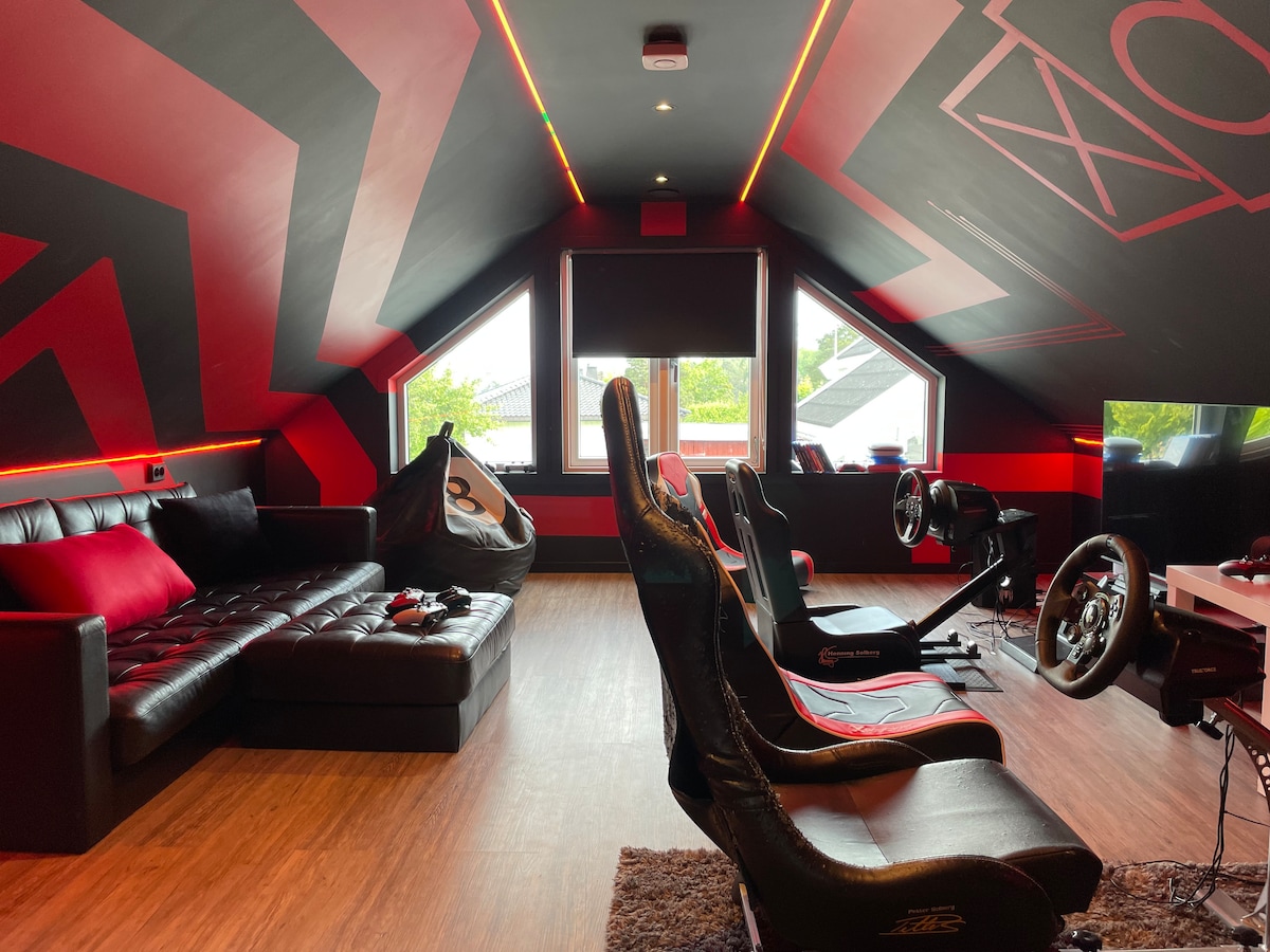 An entertainment room features a black sectional sofa and two racing-style gaming chairs, positioned toward a large screen. Bold red and black geometric designs adorn the sloped ceiling, while large windows provide natural light, framed by dark curtains. A gaming setup is visible, enhancing the space.