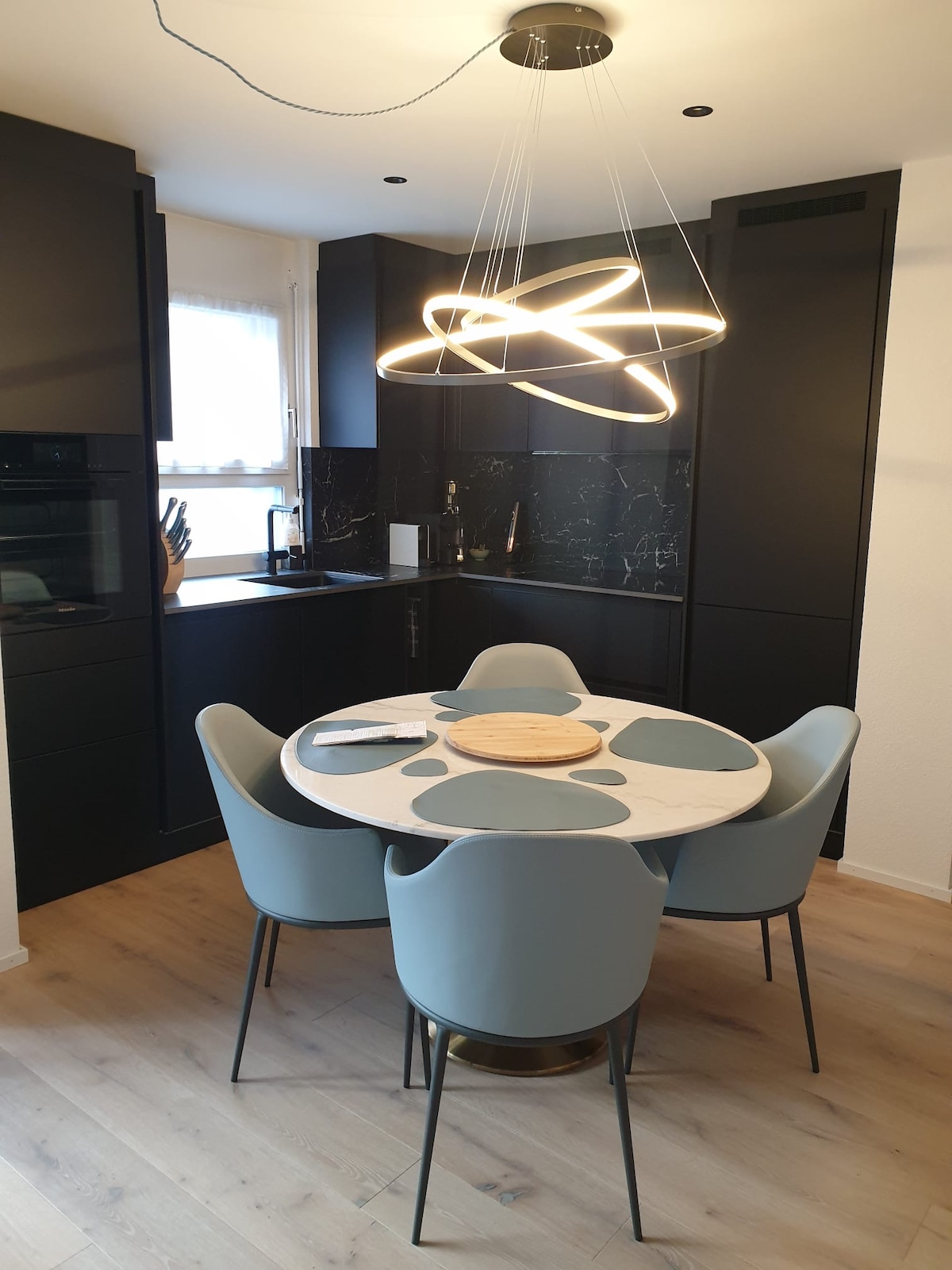 A modern kitchen area is showcased with sleek black cabinetry and a marble dining table surrounded by four light grey chairs. A unique pendant light hangs above the table, providing a bright focal point. Natural light filters in through a nearby window.