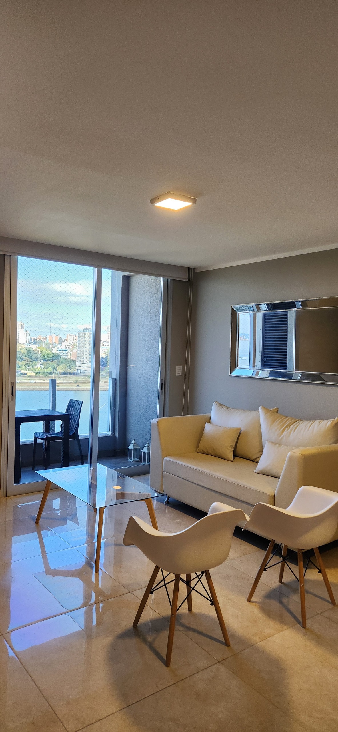 A modern living area is presented with a light-toned sofa and two white chairs positioned around a glass coffee table. Natural light brightens the space, with large glass doors leading to a view of the cityscape and water. A wall-mounted mirror enhances the room's openness.