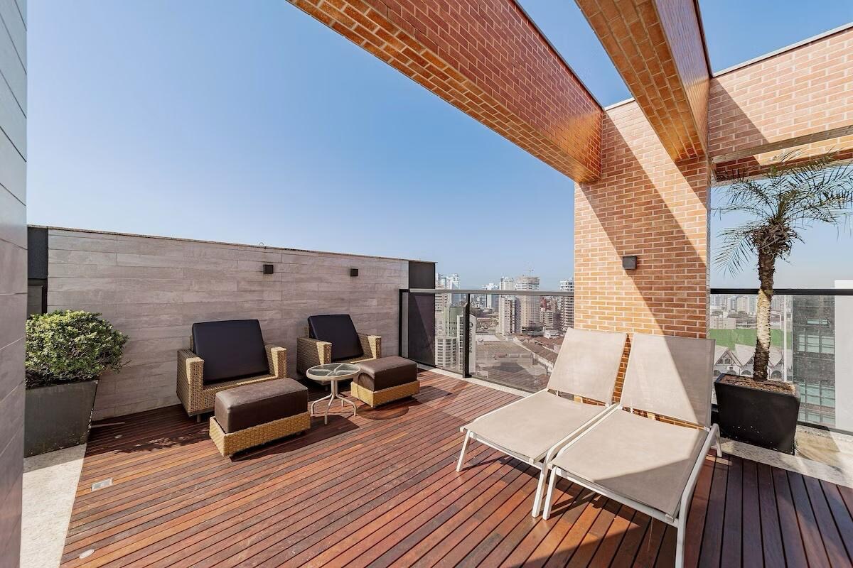 An outdoor terrace is presented featuring two lounge chairs and a small table on wooden decking. Potted greenery adds a touch of nature, while a partially obstructed city skyline can be seen in the background under clear blue skies.