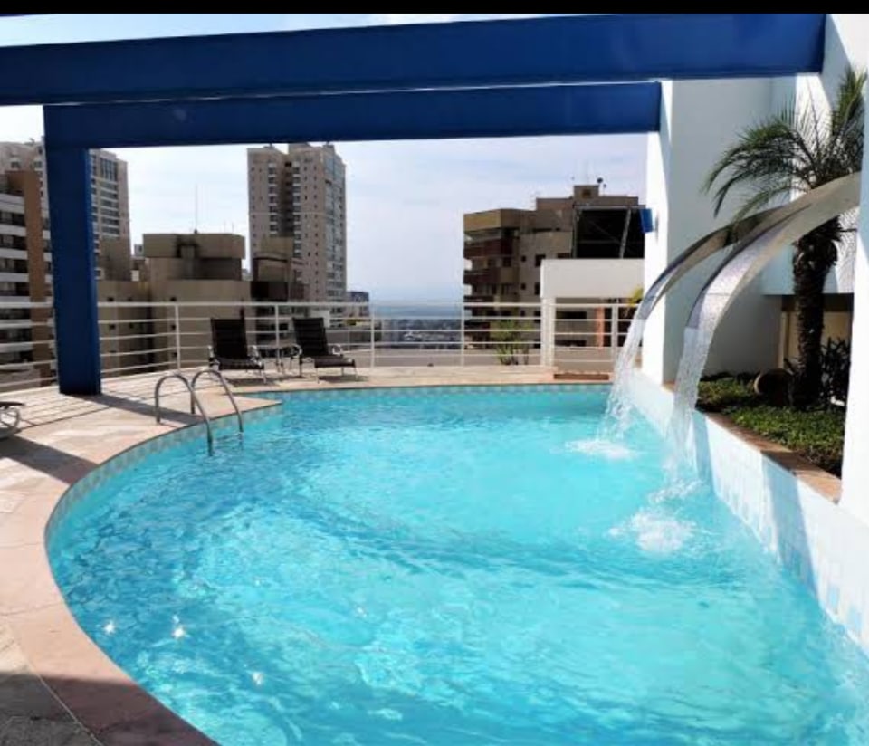A rooftop pool area features clear blue water and a cascading waterfall, surrounded by a paved deck. Lounge chairs are positioned nearby, offering a space for relaxation. The skyline of the city is visible in the background, enhancing the tranquil setting.