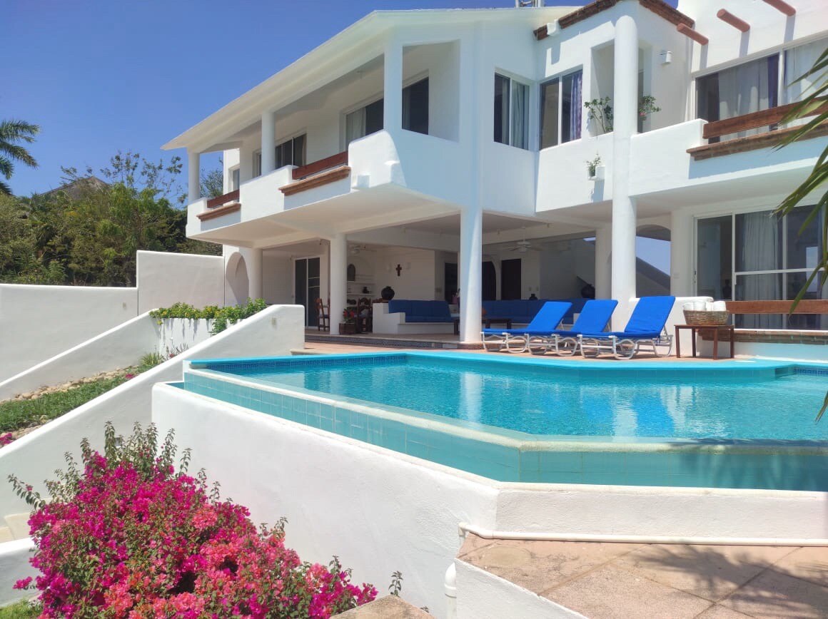 A modern villa is depicted, featuring a sparkling swimming pool surrounded by colorful flowering plants. Sun loungers in bright blue line the poolside. The structure boasts multiple levels, with large windows that invite natural light into the interior spaces.
