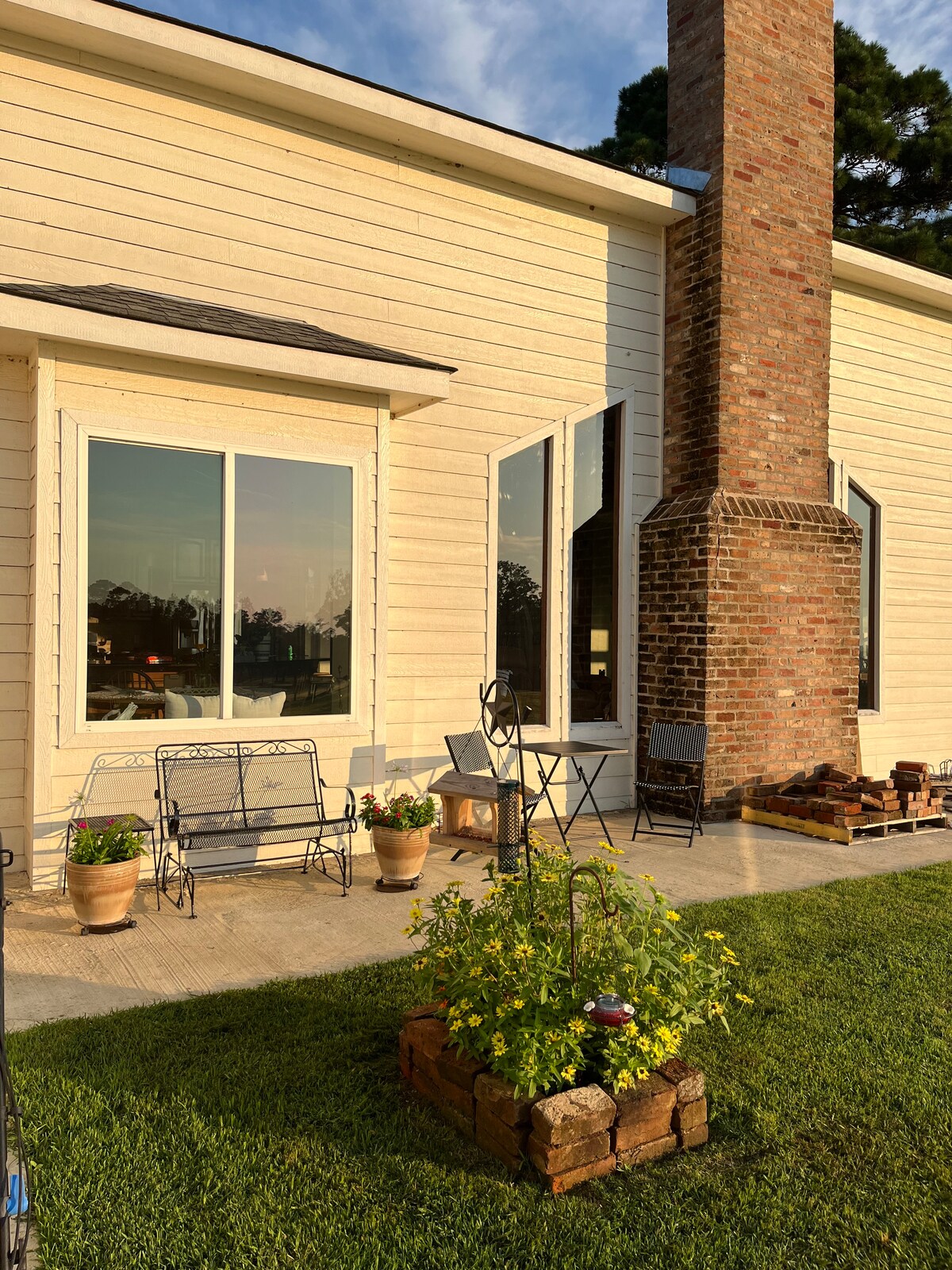 A charming outdoor patio area is featured, complete with a small table and several decorative chairs. Potted plants add a touch of greenery, while a stone border surrounds a blooming flower bed. Natural light catches the brick chimney, enhancing the cozy setting.