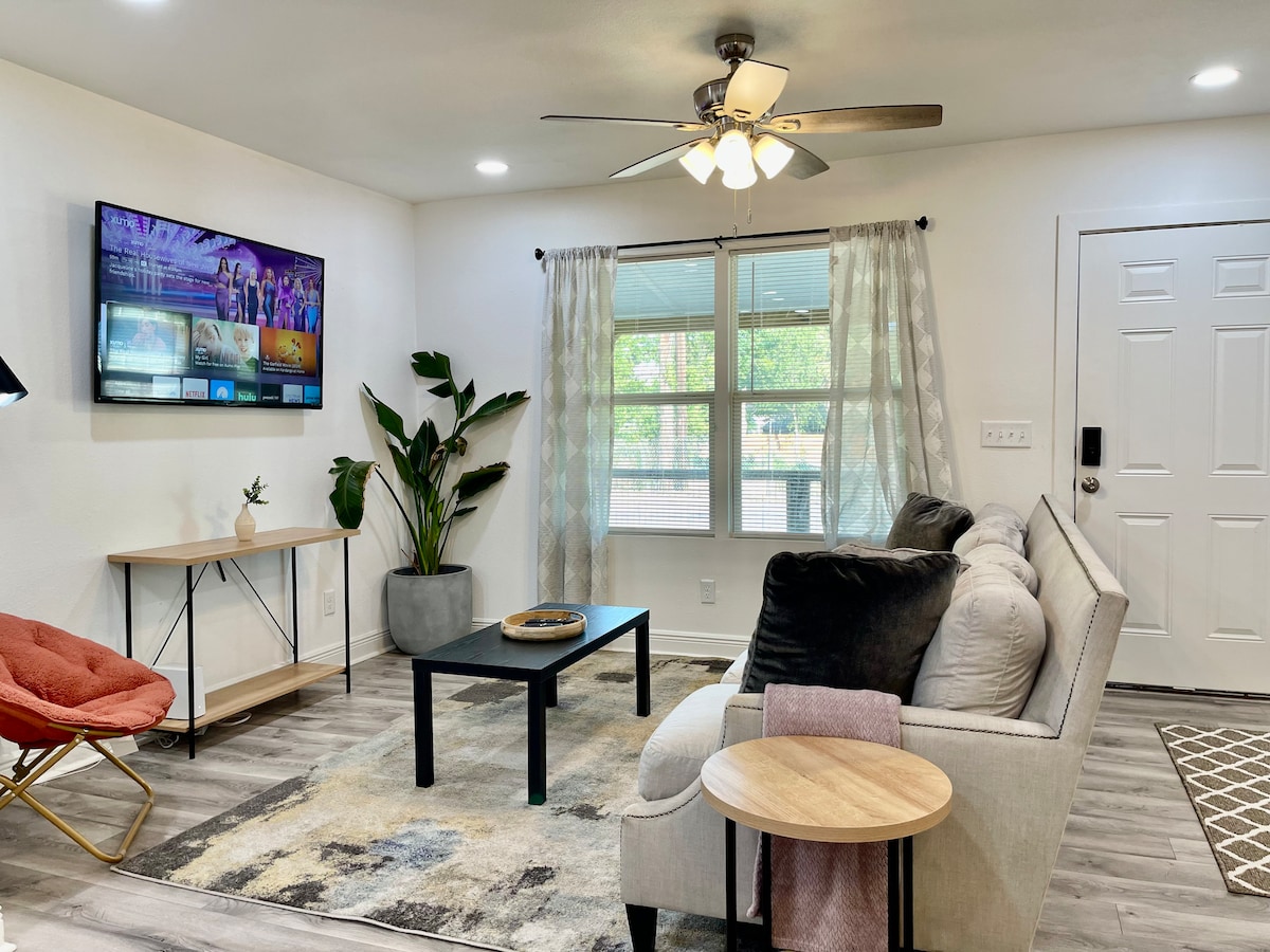 An open-concept living area is shown, featuring a comfortable sectional sofa and a black coffee table on a textured rug. Large windows allow for natural light, while a ceiling fan and wall-mounted TV enhance the inviting atmosphere. A potted plant adds a touch of greenery.
