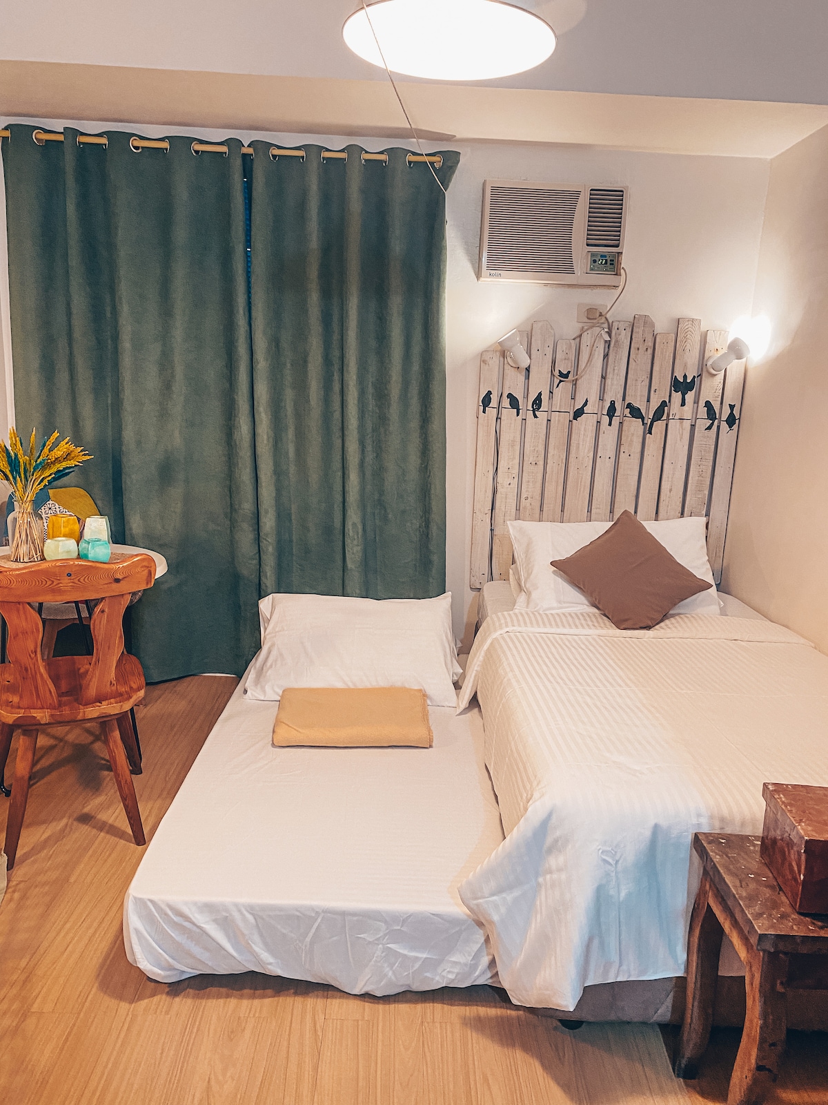 A neatly arranged studio features a spacious bed and a separate floor mattress, both dressed in white linens. A round wooden table with colorful decorative bottles sits nearby, while a set of green curtains frame a window, adding a touch of color to the space.