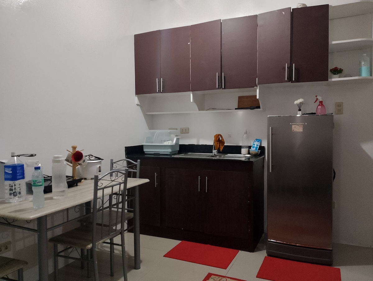 The kitchen area features dark wood cabinets and a stainless steel refrigerator. A small dining table is set with four chairs. Counter space is available for meal preparation, with a sink and a compact dish rack visible.