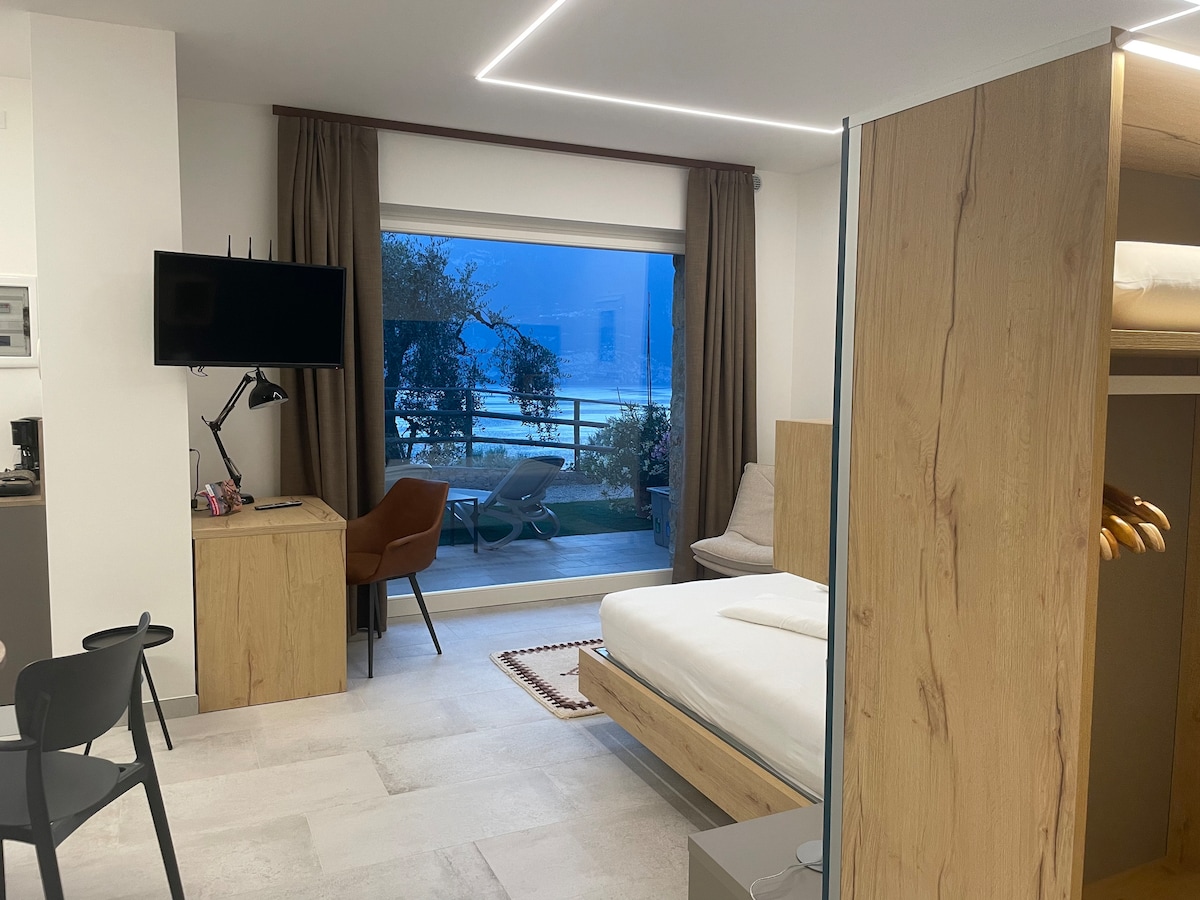 A modern studio features a spacious bed with a light wooden frame. A large window offers a view of the lake, complemented by a cozy seating area with lounge chairs on the terrace. Soft lighting and neutral tones create a calm environment.