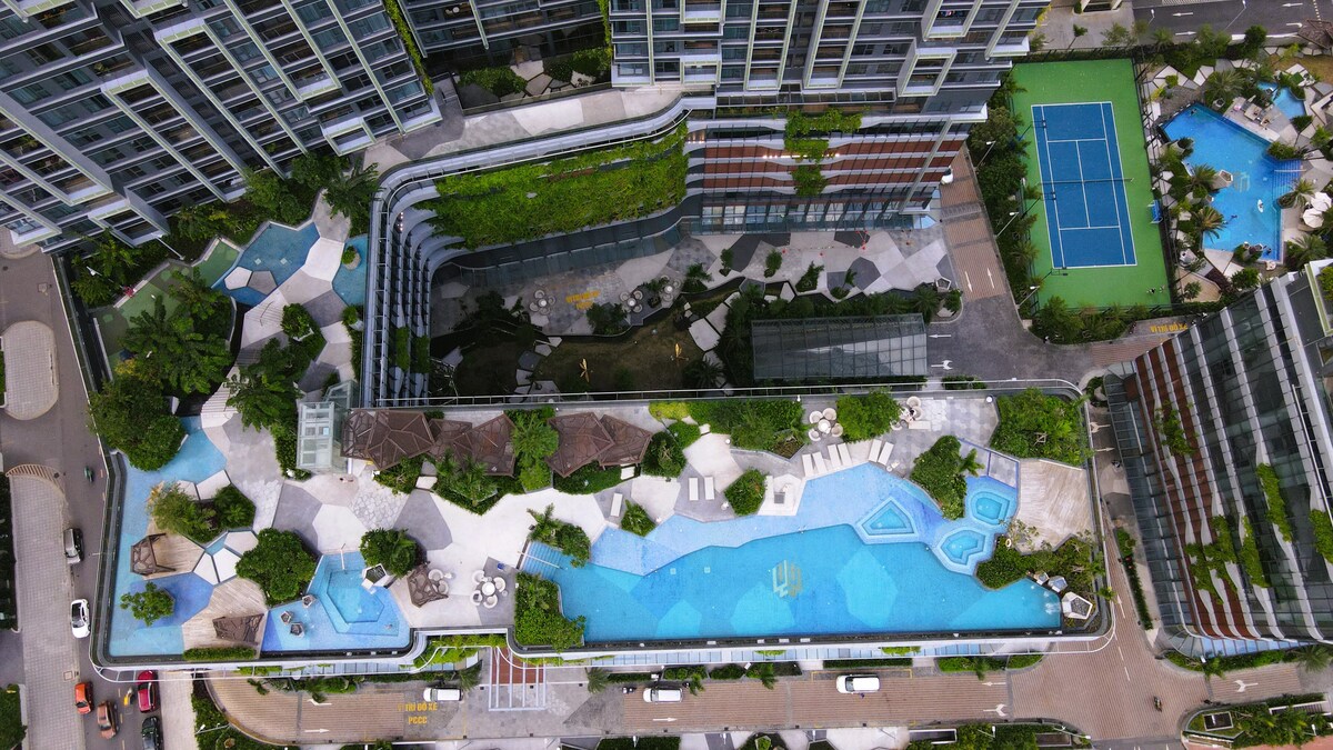 An aerial view of the condominium complex showcases a multi-level pool area surrounded by lush greenery. The salty water infinity pool extends across the rooftop, complemented by lounge areas and a tennis court visible in the background. The overall layout highlights a harmonious blend of water features and landscaped spaces.