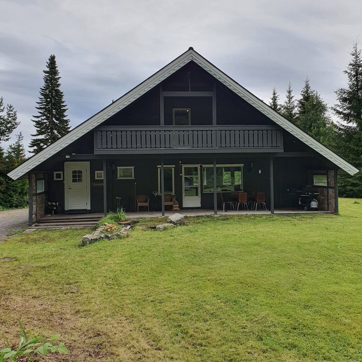 The exterior of a two-story house is showcased, featuring a sloped roof and dark wooden siding. A spacious front lawn is visible with a pathway leading to the entrance. Nearby seating is arranged on the porch, and a barbecue grill is positioned to the side.
