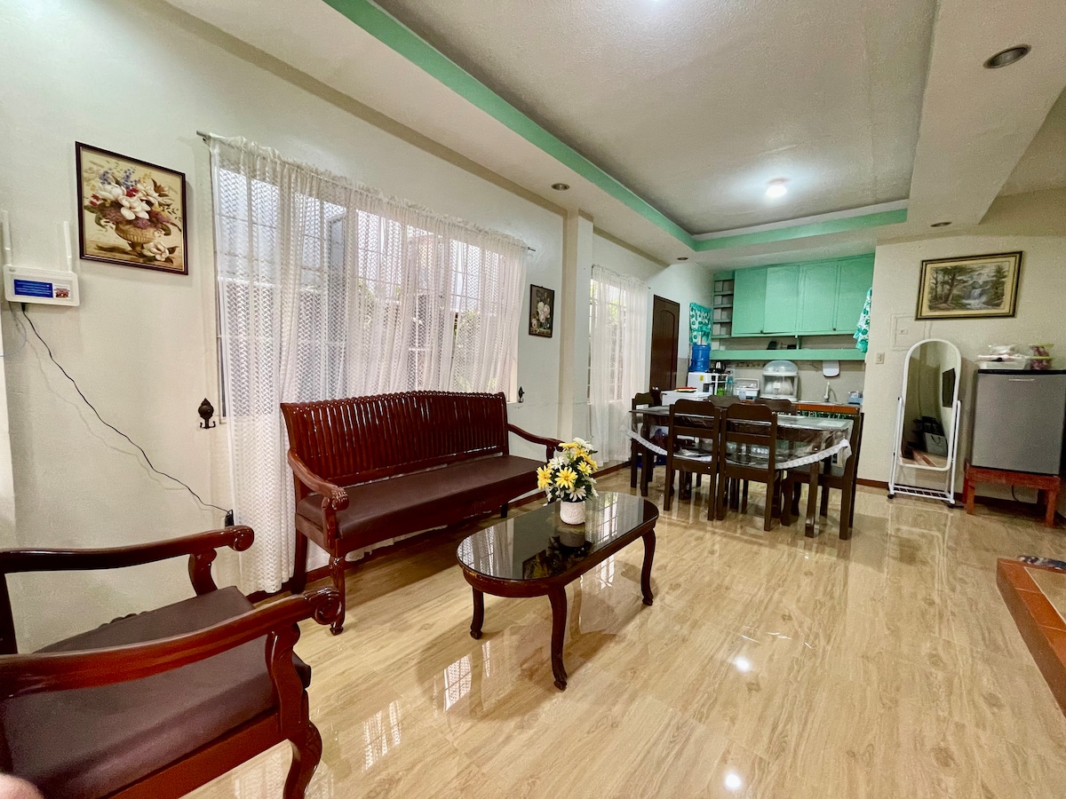 The open living and dining area features hardwood seating, a glass coffee table, and a spacious dining set. Natural light is provided by large windows adorned with sheer curtains, while a functional kitchen can be seen in the background, showcasing green cabinetry.