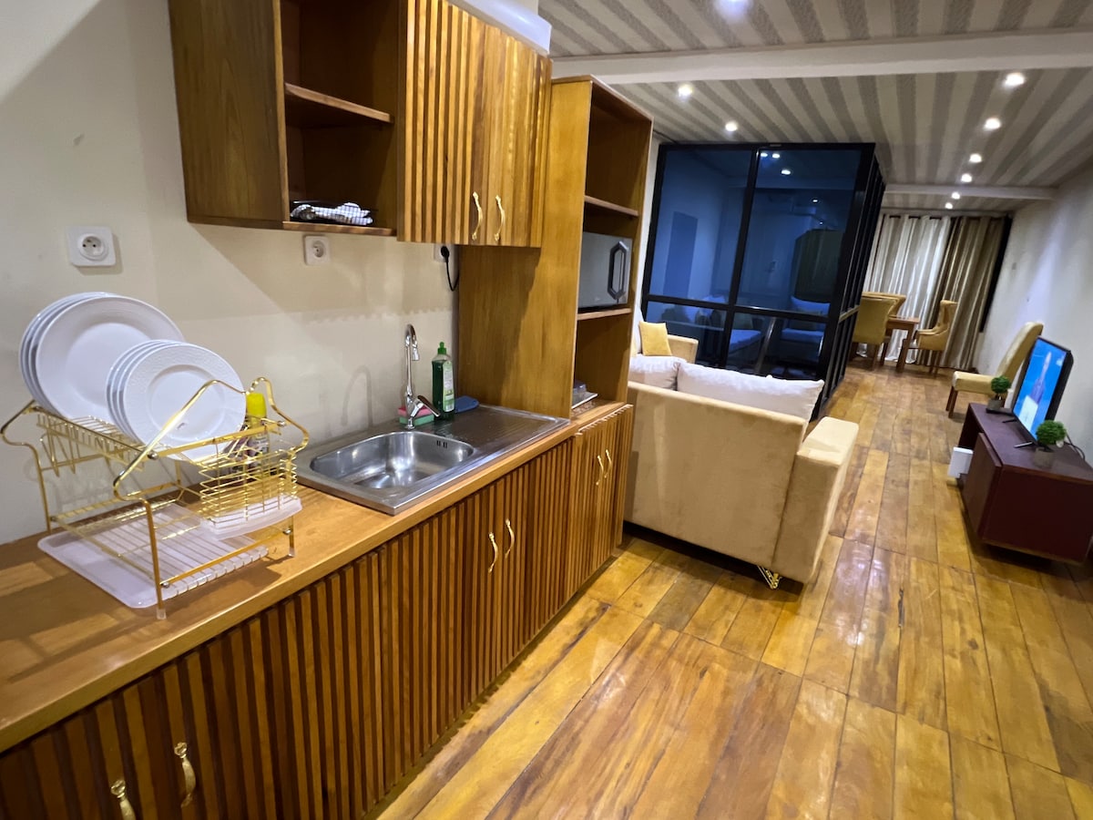 A well-organized kitchen space showcases wooden cabinetry and a stainless steel sink. A dish drying rack is positioned beside the sink, and a refrigerator is integrated into the cabinetry. Adjacent, a cozy living area can be seen with light-colored furniture and a television.
