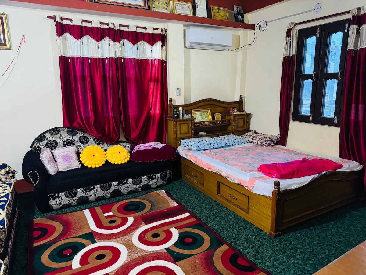 An inviting room features a wooden bed with bedding and a decorative couch adorned with colorful cushions. Rich red curtains frame the windows, allowing natural light to enhance the ambiance. A patterned rug adds texture to the floor, complementing the room's warm tones.