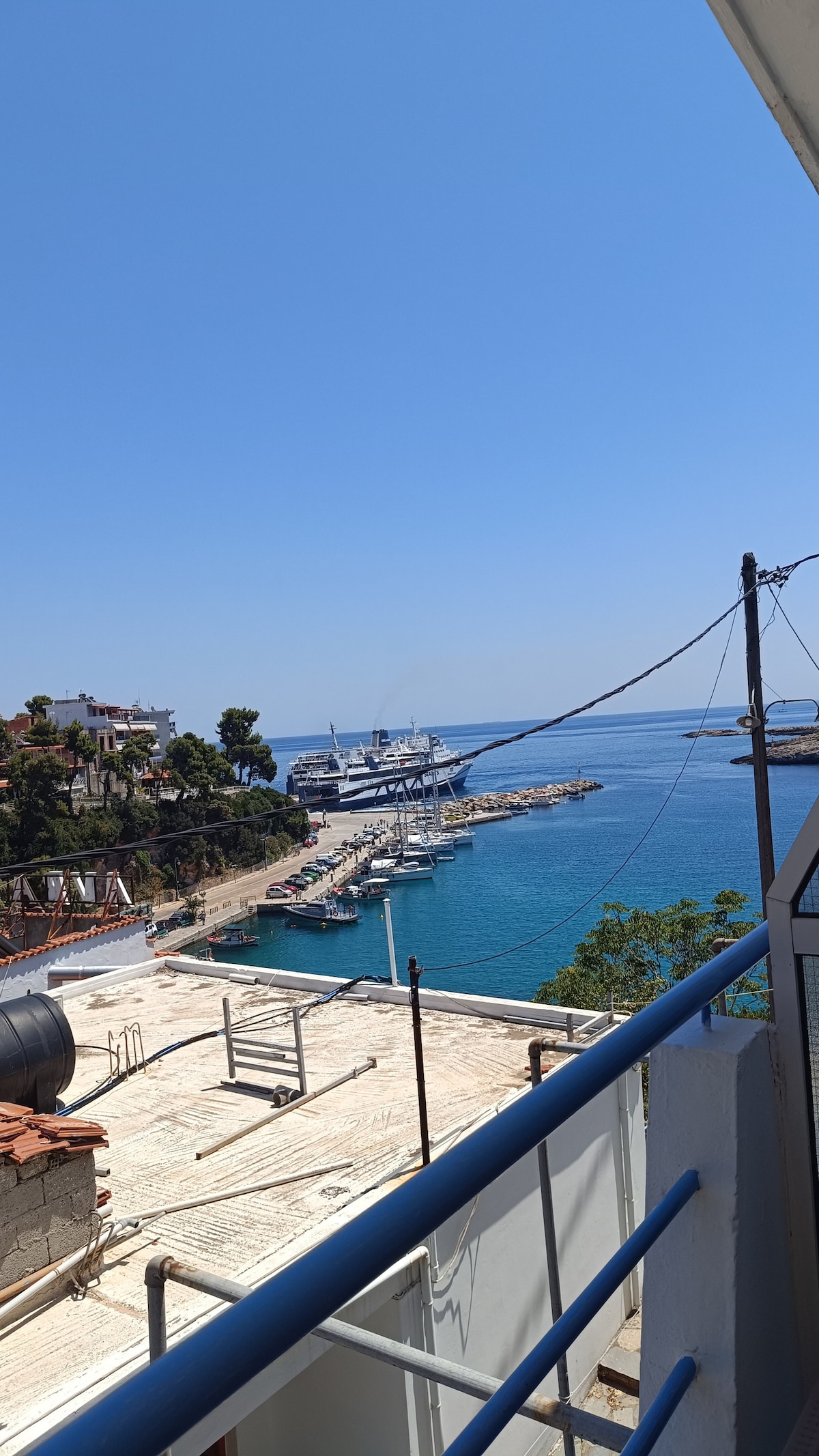 A clear view of the harbor can be seen from the balcony, showcasing the water's vibrant blue hue. The marina is lined with several boats, while the shoreline features a mix of greenery and buildings in the background, under a bright, cloudless sky.
