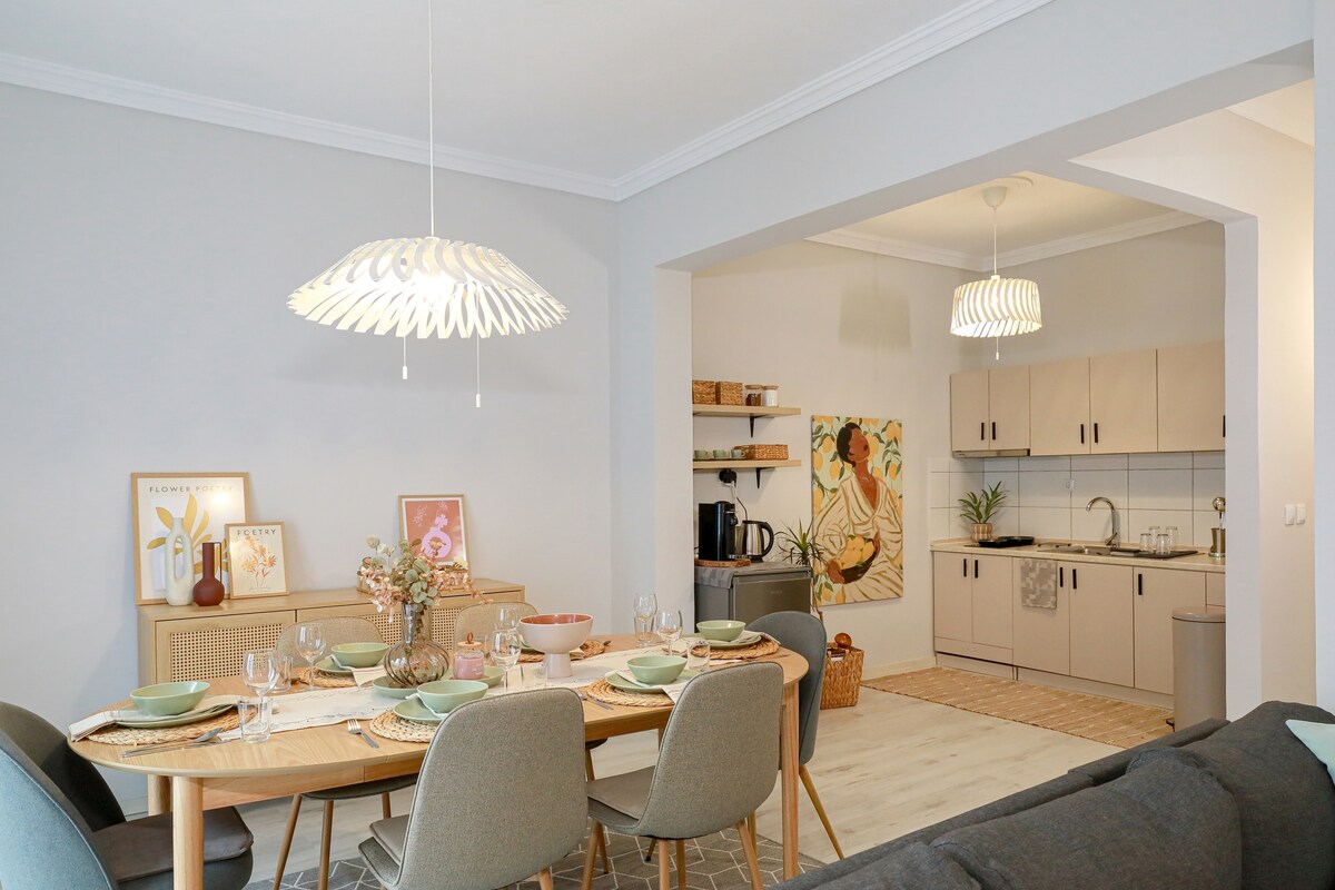 A spacious dining area features a wooden table set for a meal, with grey upholstered chairs surrounding it. Two modern pendant lights hang above, illuminating the table. The kitchen area is equipped with cabinetry and modern appliances, providing a welcoming open-plan layout.