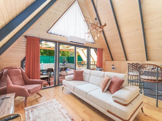 An A-frame interior is showcased with high wooden ceilings and large windows allowing ample natural light. A modern sectional sofa is complemented by textured cushions, while a plush armchair adds to the seating options. Outdoor views can be seen through the glass doors leading to a deck.