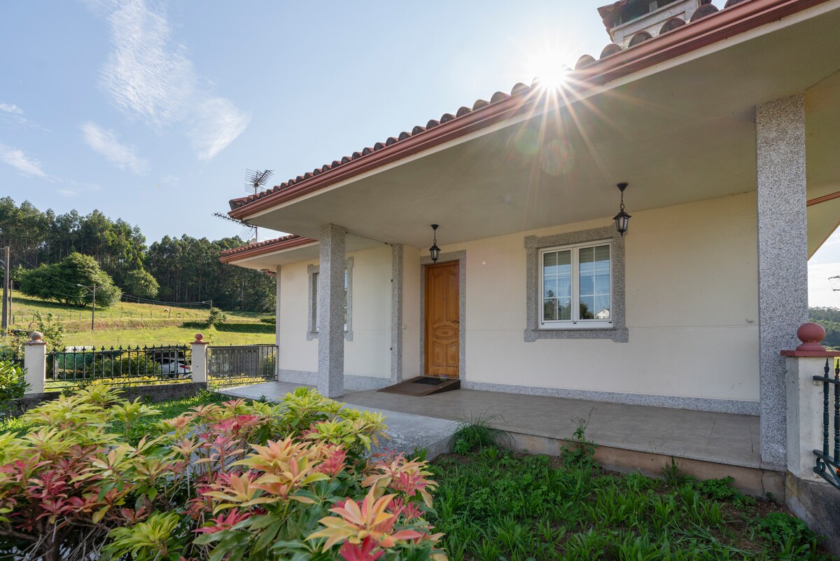 The inviting exterior of the house is captured, featuring a covered entryway framed by decorative columns. A well-maintained garden with vibrant foliage is visible, along with a large window that allows natural light to brighten the interior space. Scenic green hills are seen in the background.
