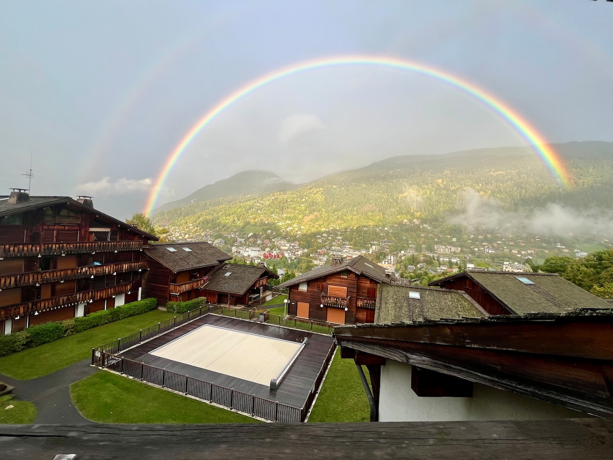 A panoramic view captures a vibrant rainbow arching over the mountainous landscape. Below, wooden structures are framed by lush greenery, with a large flat area that can be utilized for various activities. The scene conveys a peaceful connection to nature.