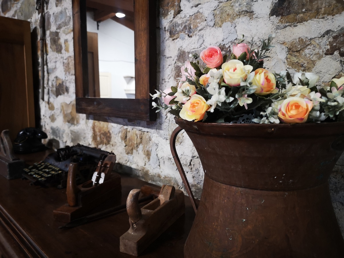 A rustic decor arrangement features a vintage metal pitcher filled with artificial roses and greenery. A mirror reflects the stone wall, while wooden tools and an old-fashioned typewriter are showcased on a solid wooden surface.