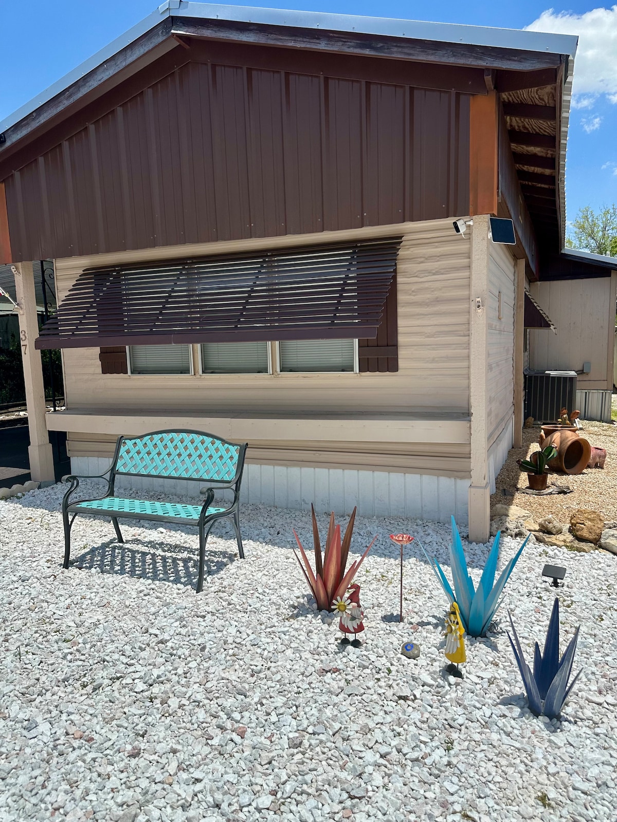 The exterior of the mobile home features a covered porch with a turquoise bench positioned on gravel. Decorative plants are arranged in the foreground, showcasing various colors and forms. The building has a combination of brown and cream siding, with windows adorned by wooden slats.