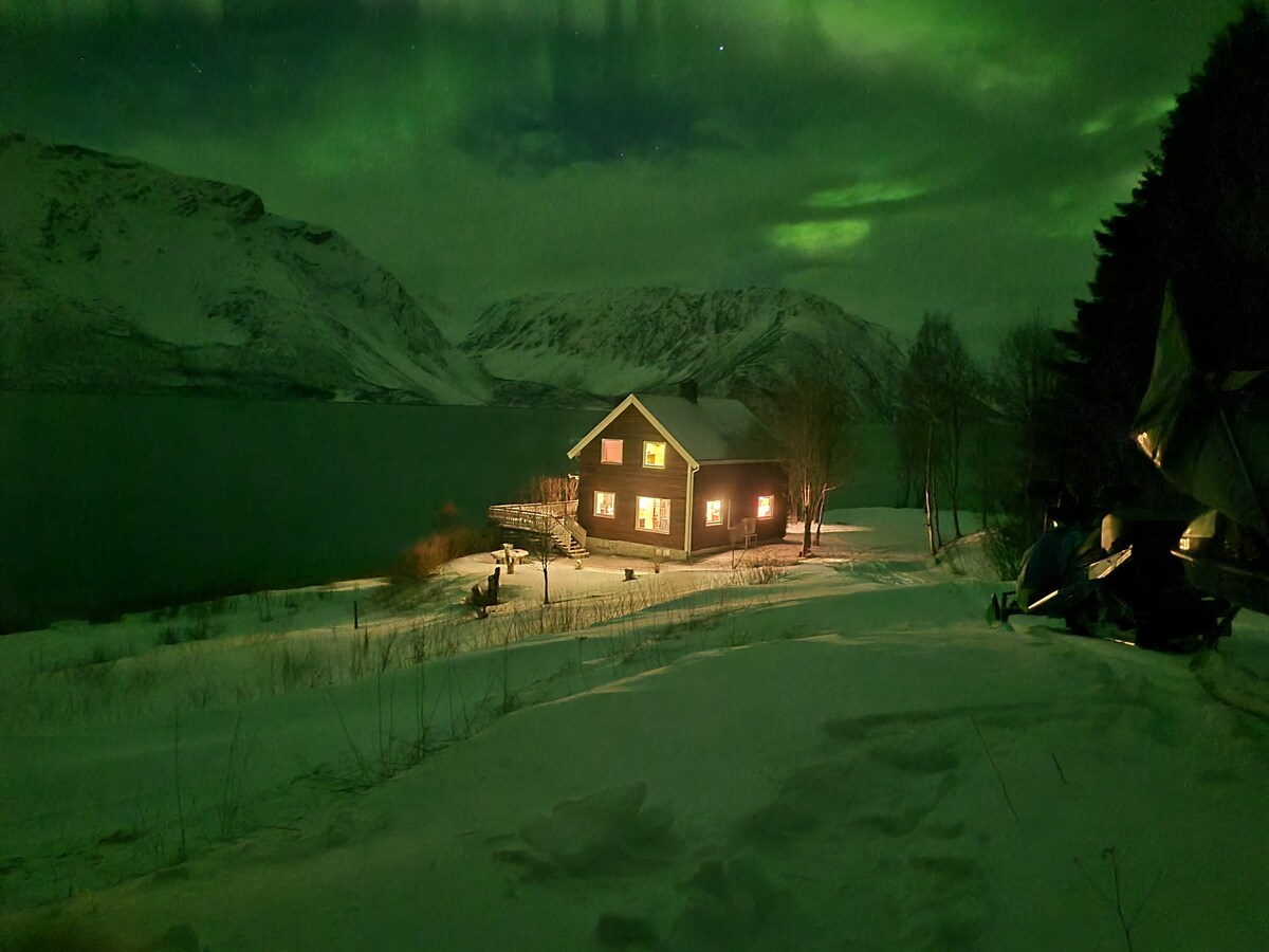 A two-story house is illuminated by warm lights against a backdrop of snowy terrain and a tranquil body of water. The northern lights dance in the night sky above distant mountains, adding a serene glow to the winter landscape.