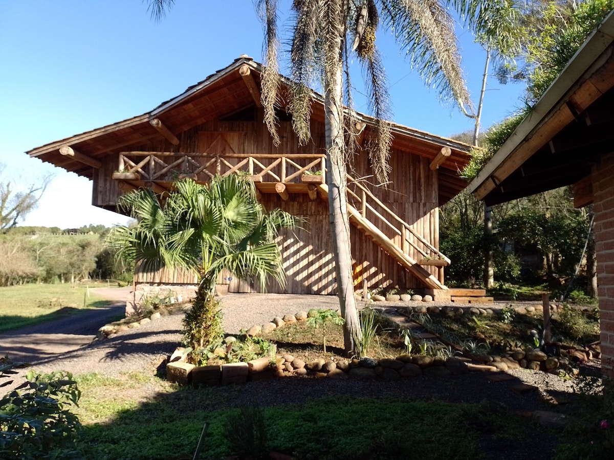A rustic wooden structure features a large balcony supported by sturdy beams, surrounded by lush greenery. A palm tree and well-maintained garden beds line the pathway leading to the entrance, enhancing the serene outdoor setting.