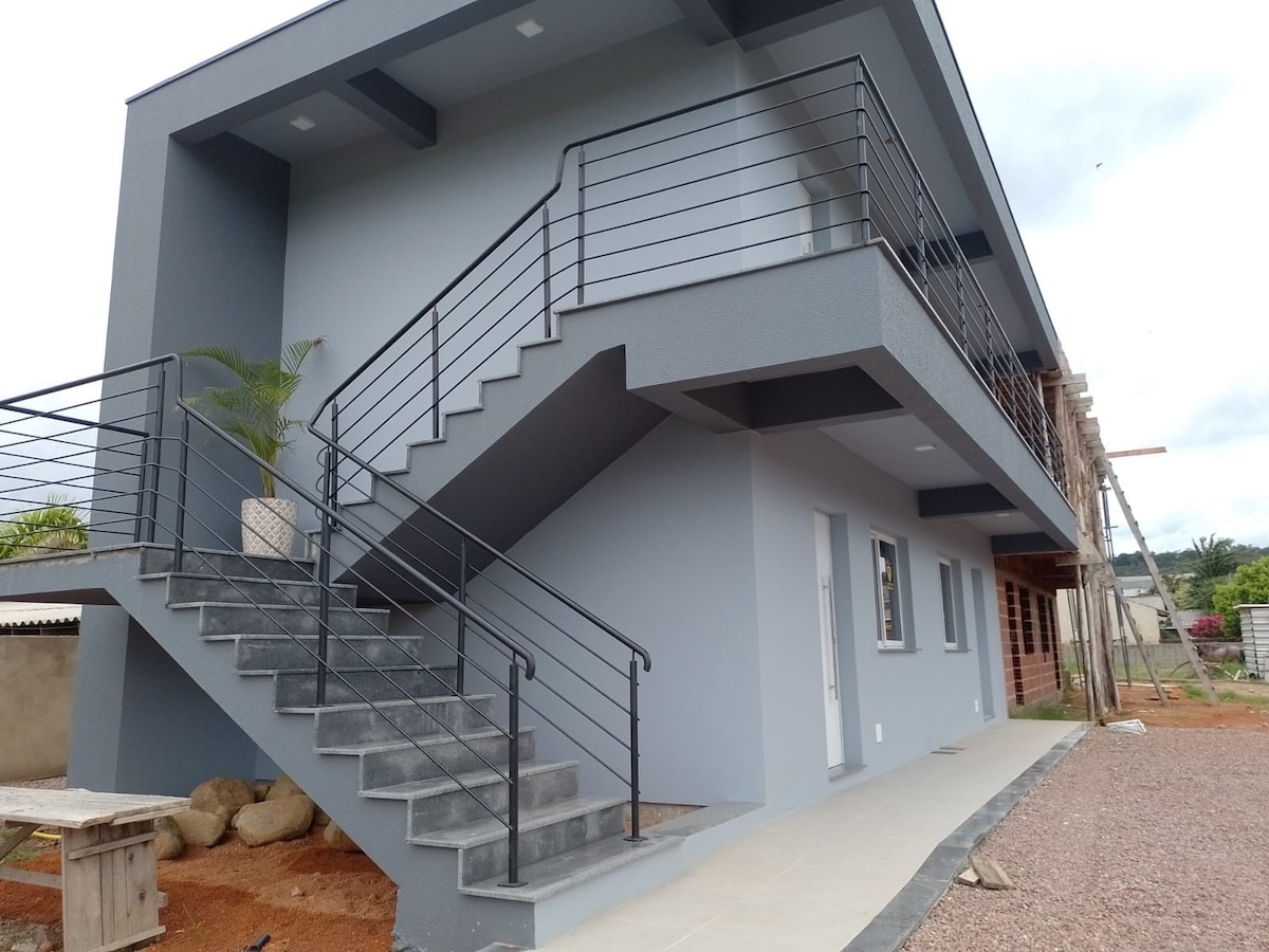 A modern two-story building is featured, displaying a sleek staircase with black railing leading to the second level. The exterior is painted in a calming gray tone, enhanced by a planted container on the landing. A pathway of tiles extends alongside the building.