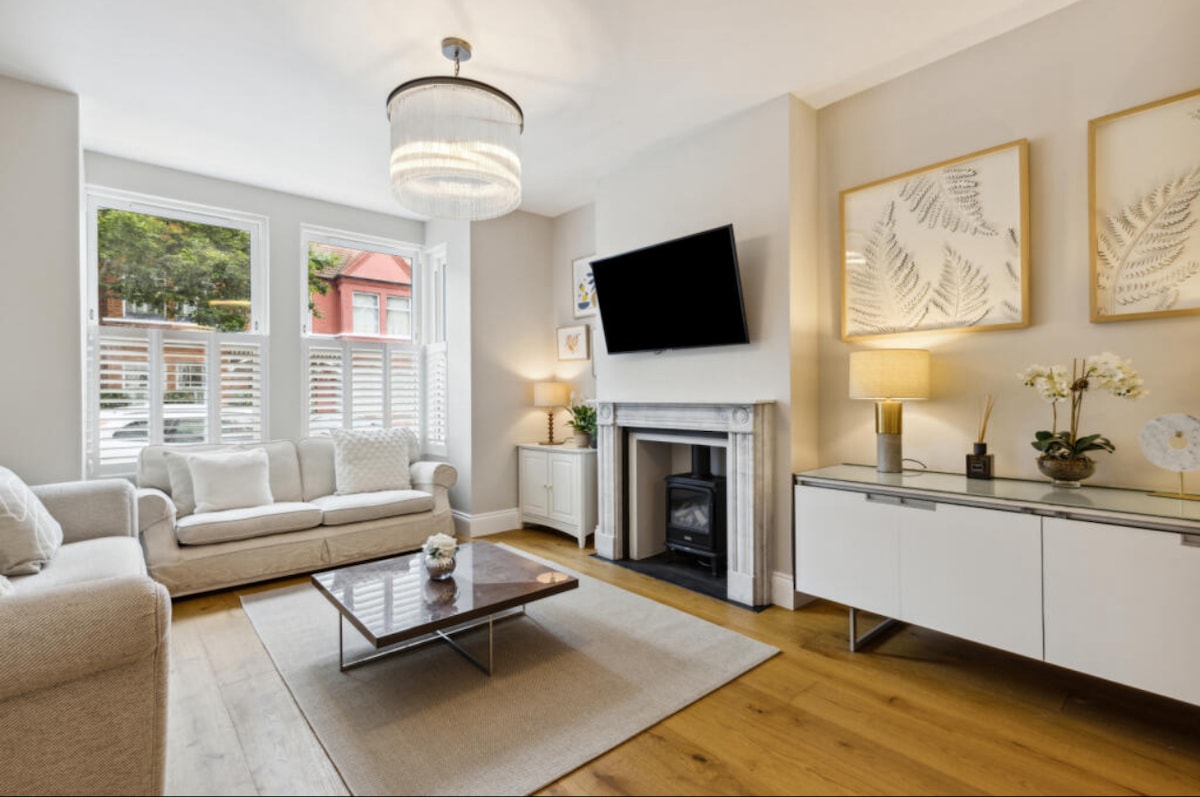 A bright living room is featured with large windows allowing natural light. A stylish fireplace is framed by light-colored walls, complemented by a modern flat-screen TV. A light-colored sofa and a low coffee table occupy the space, alongside decorative wall art and a console table.