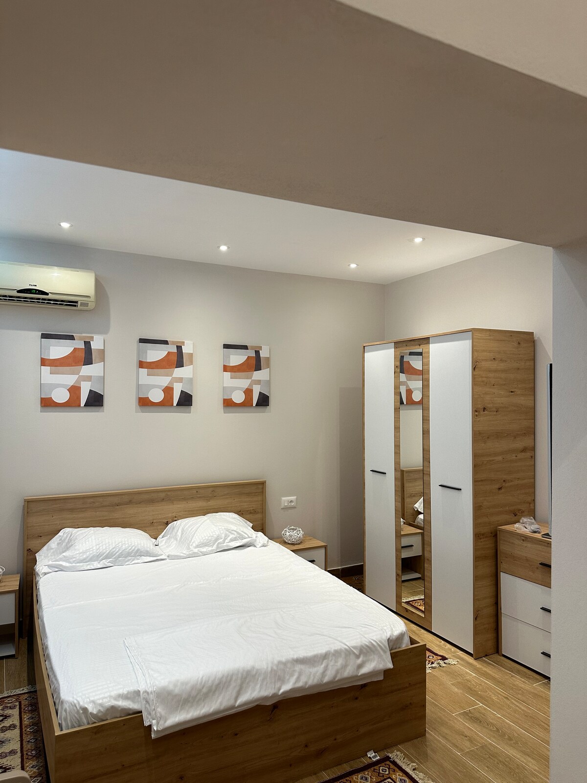A cozy sleeping area features a spacious bed with white bedding. Above the bed, three abstract prints adorn the wall. A wardrobe with a combination of wood and white finishes stands nearby, alongside a small bedside table.