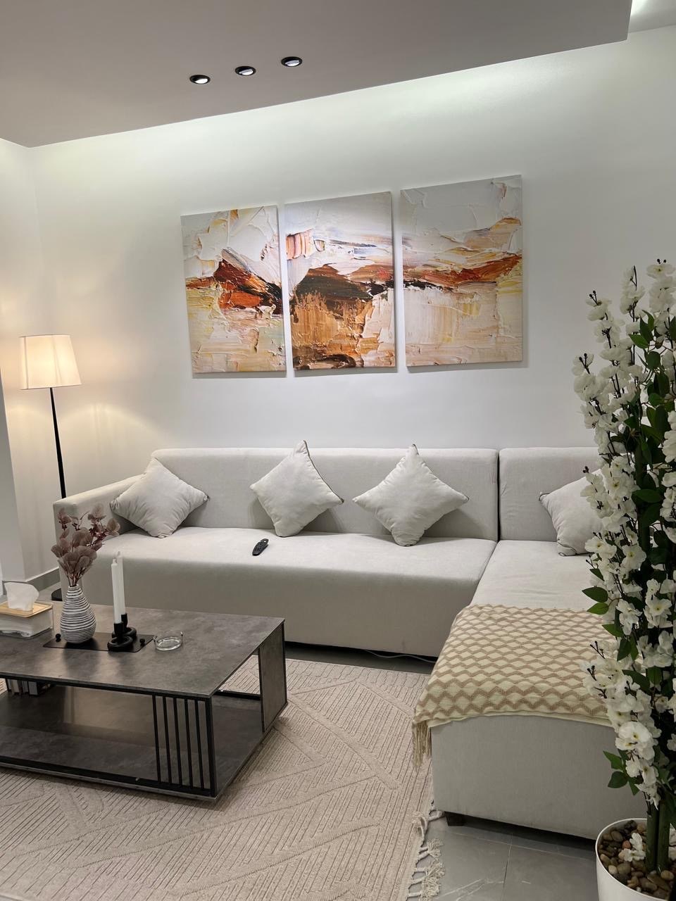 A modern living space features a light-colored sectional sofa adorned with decorative pillows. A coffee table rests in front, accompanied by a candle and decorative elements. Three art pieces hang on the wall, offering a subtle contrast. Natural light is enhanced by a floor lamp.