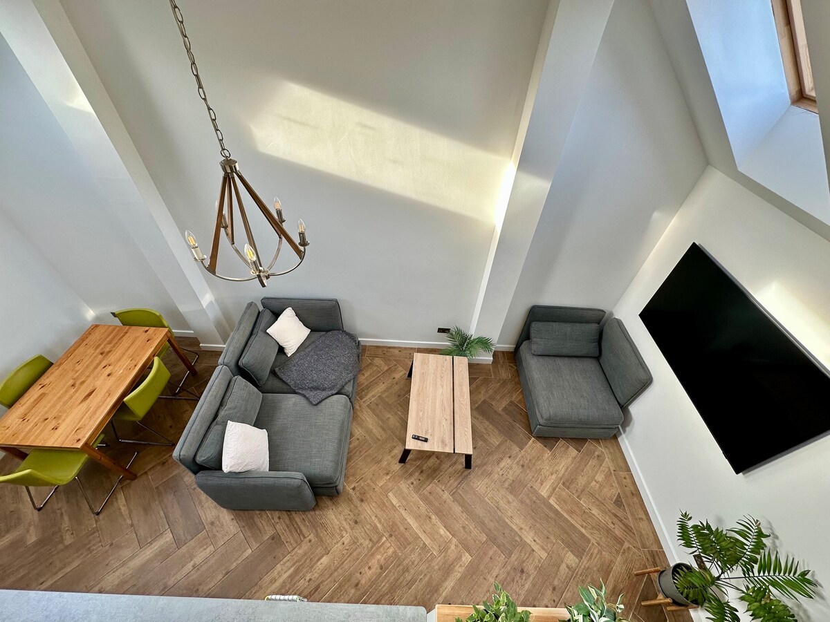 An open-plan living area features a high ceiling and modern furnishings. A spacious coffee table is centered between two gray sofas, while a wooden dining table with green chairs is positioned nearby. Natural light fills the space from the windows above.