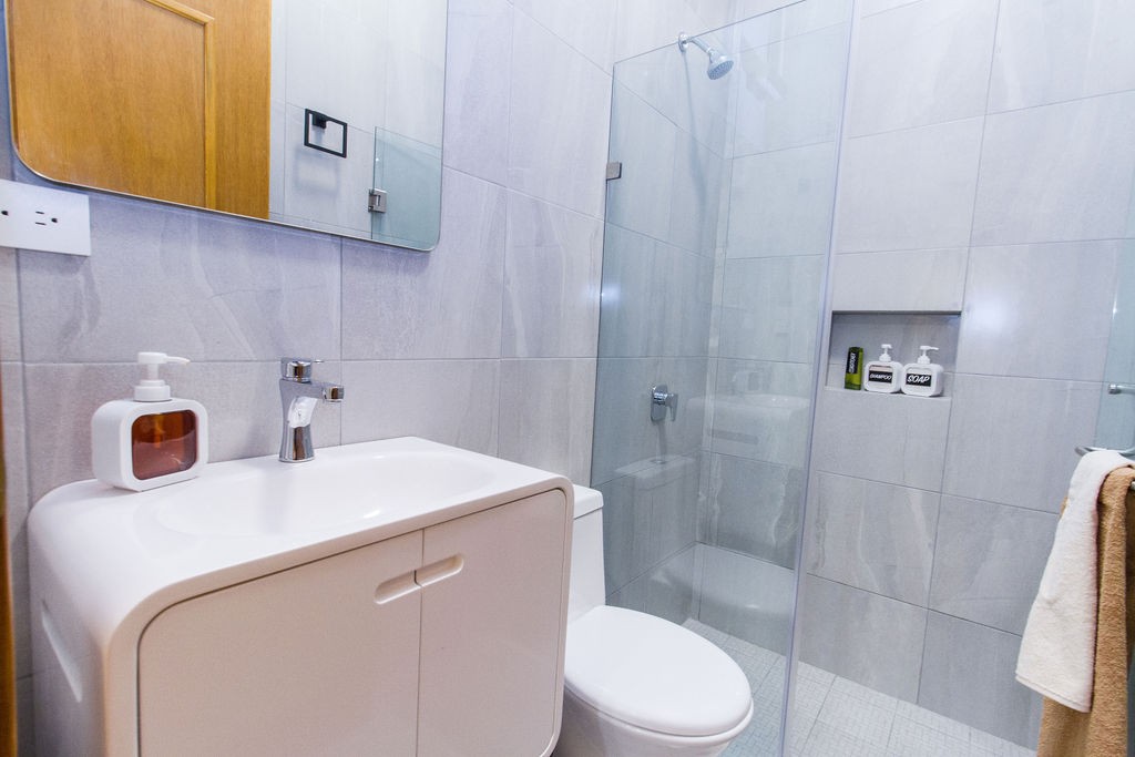 A modern bathroom is featured with a walk-in shower enclosed in clear glass. A sleek vanity with a rounded design holds a soap dispenser and other toiletries. Neutral-toned tiles complement the overall clean aesthetic, with a towel hanging nearby.