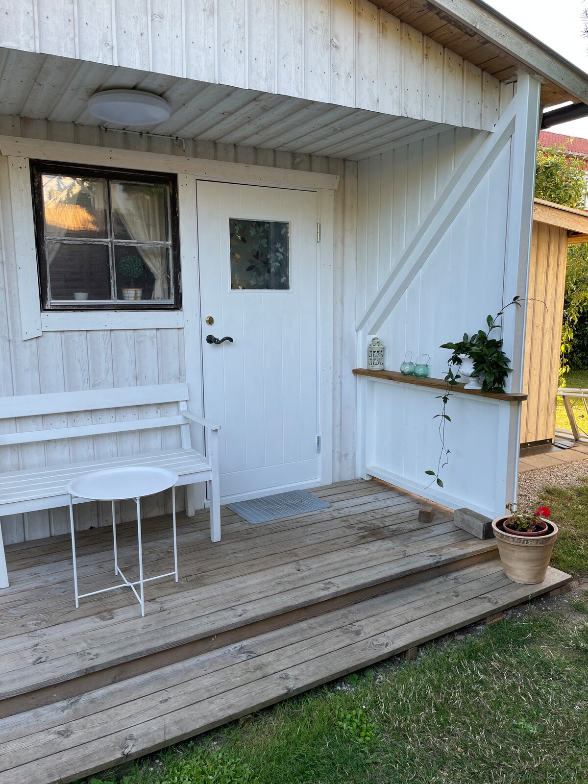 A welcoming entrance features a white door with a small window. A bench and a round table are situated on the wooden deck, accompanied by a potted plant. Light wood siding creates a bright, clean aesthetic, with grassy surroundings visible.