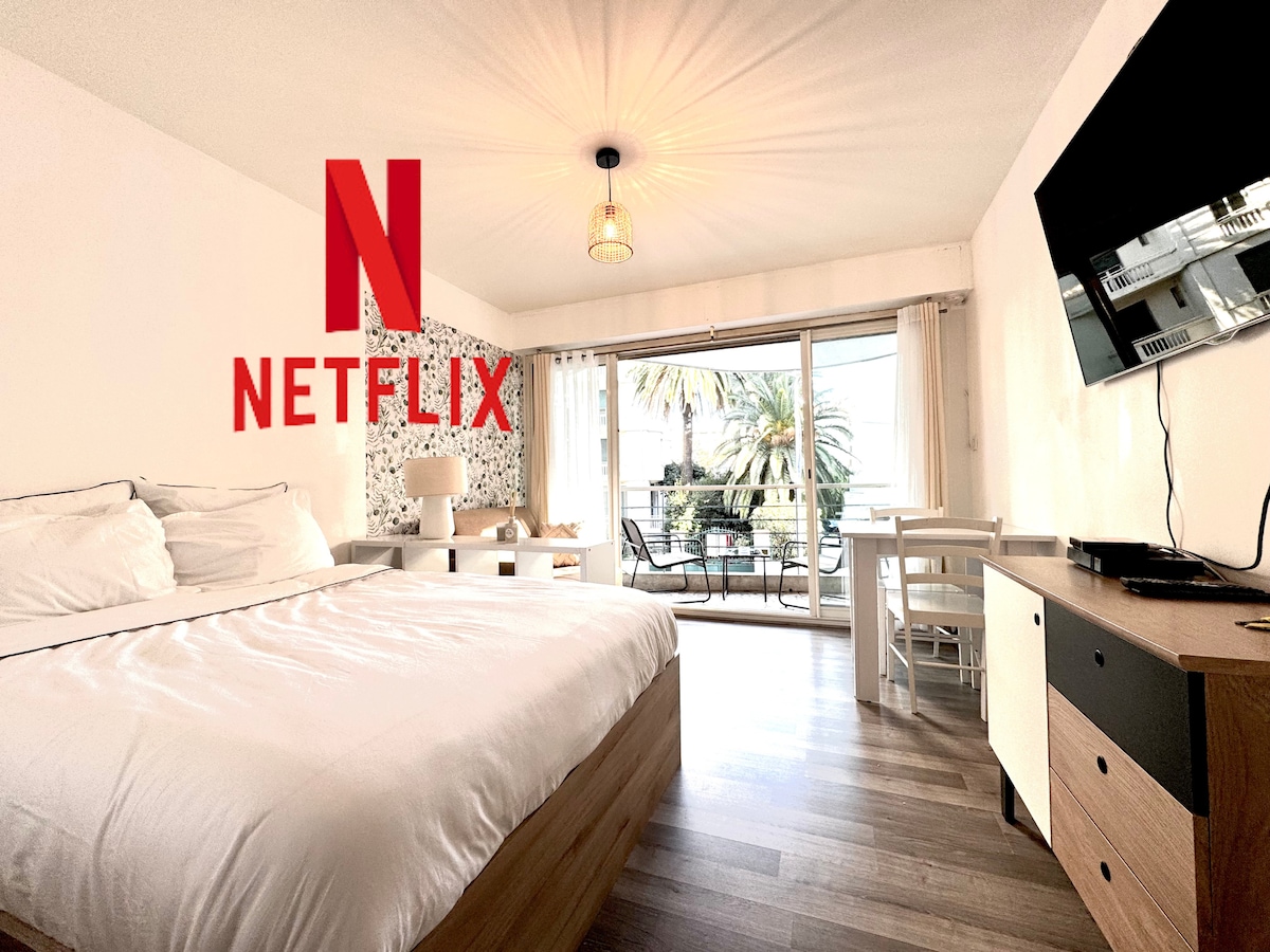 A spacious room features a large bed with crisp linens and a window showcasing outdoor views. A wall-mounted TV is positioned across from the bed, and a dining area with a table and chairs is visible. Natural light filters in through sheer curtains, enhancing the bright atmosphere.